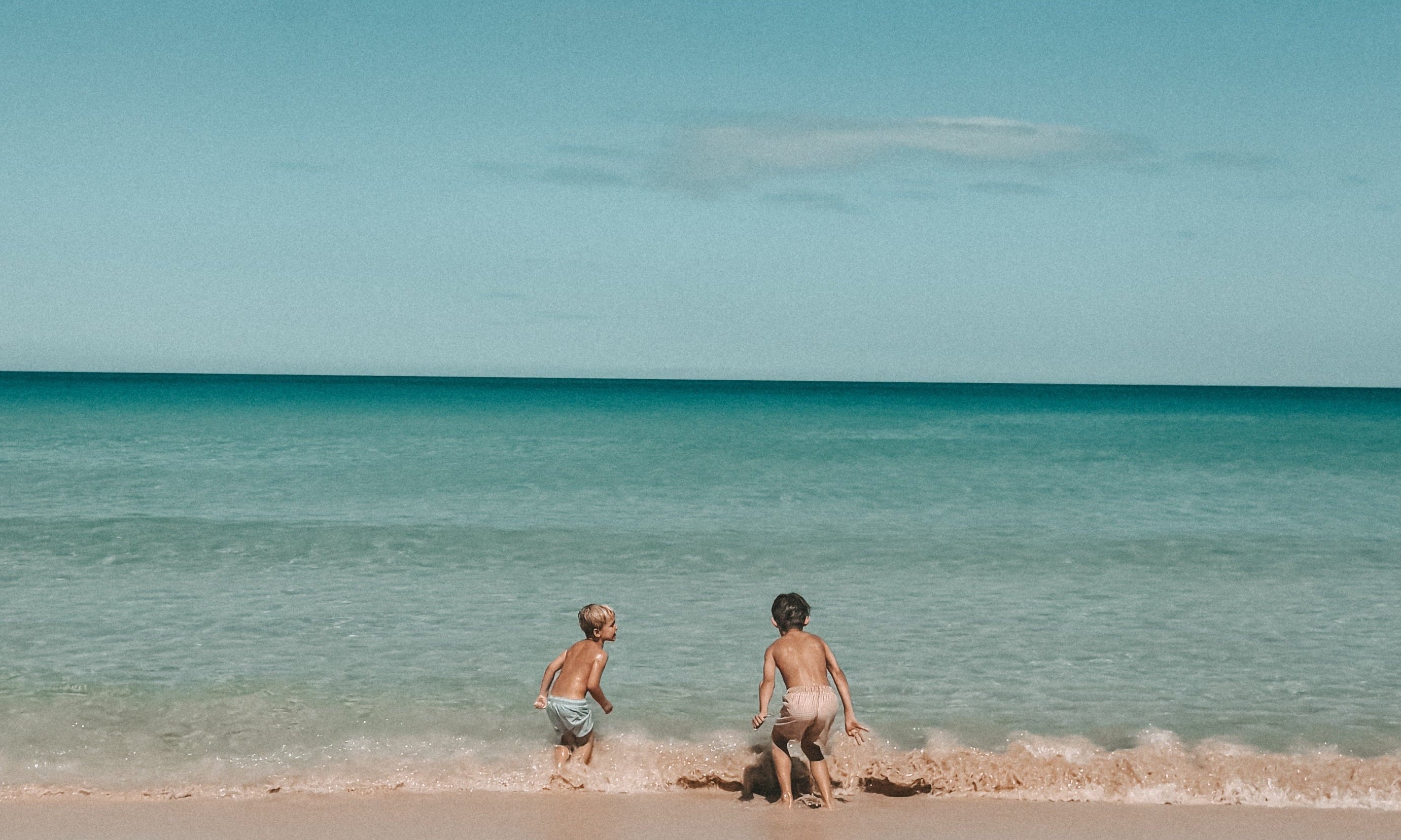 A wide beach scene with two people standing in shallow turquoise water near the shoreline under a deep blue sky with a few white clouds.