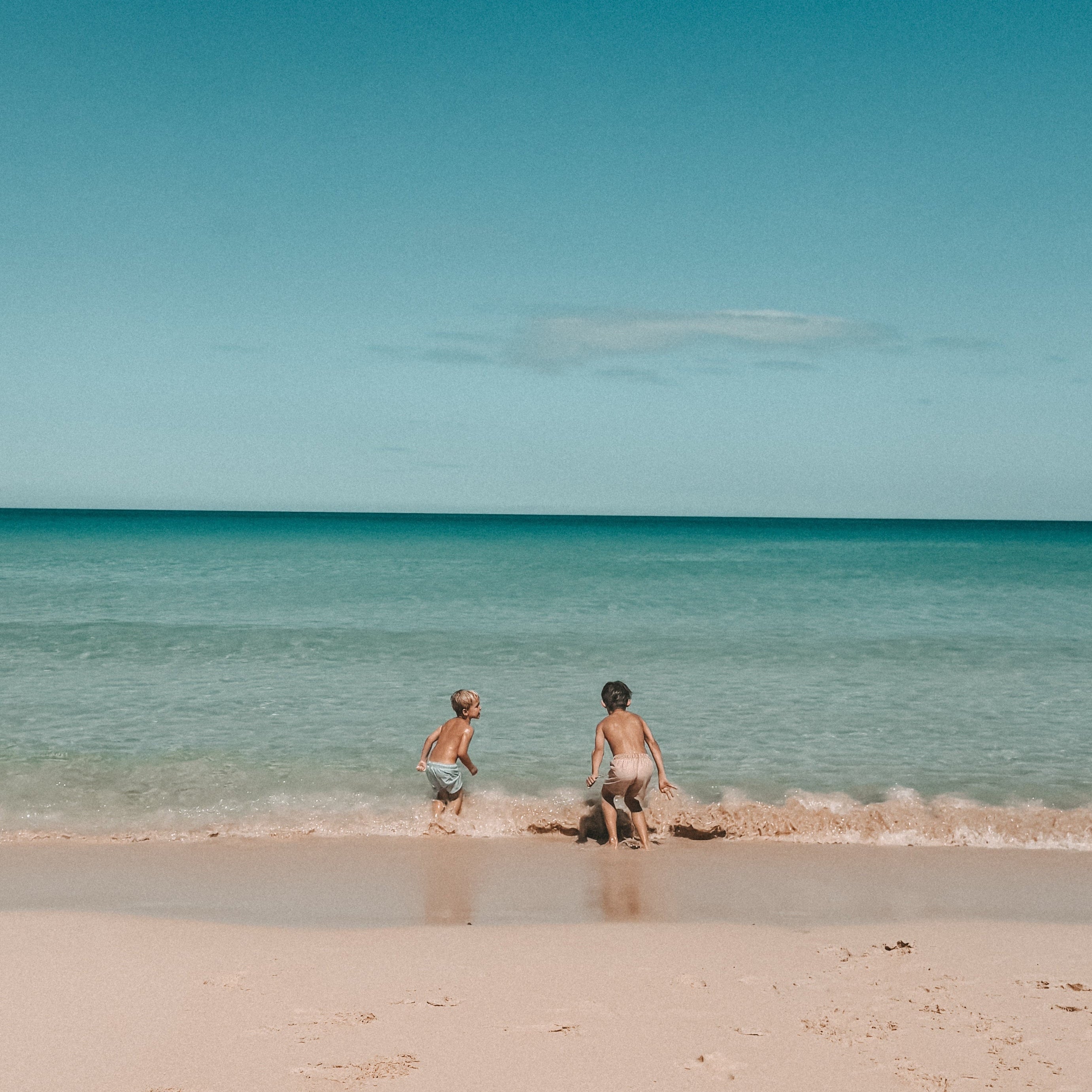 A wide beach scene with two people standing in shallow turquoise water near the shoreline under a deep blue sky with a few white clouds.
