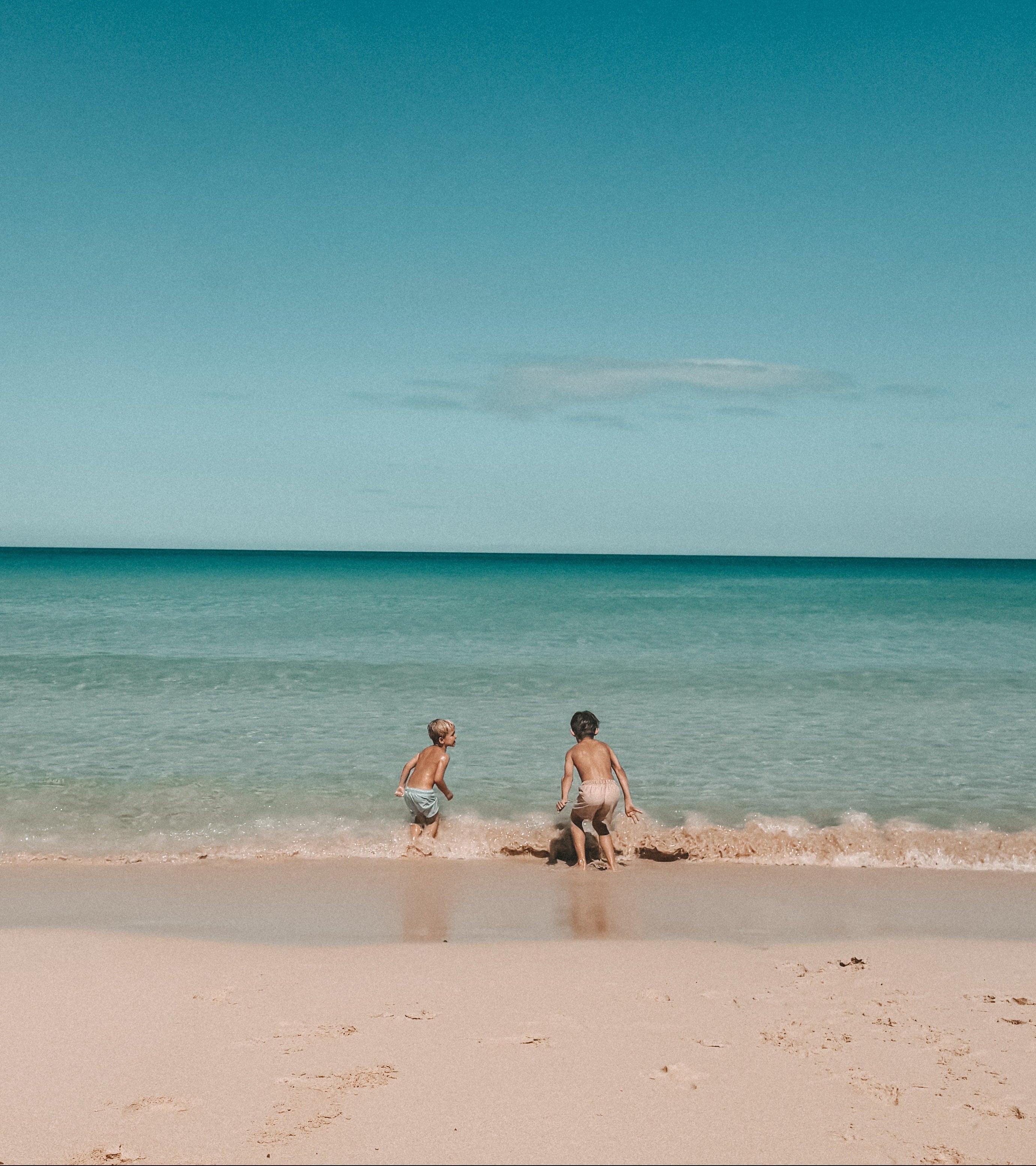 A wide beach scene with two people standing in shallow turquoise water near the shoreline under a deep blue sky with a few white clouds.