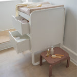 Baby changing table with open drawer and small table with items on a wooden floor.