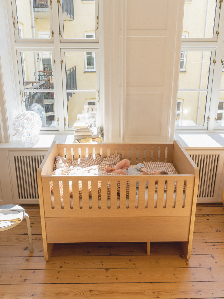 Wooden crib with a baby inside in a room with large windows.
