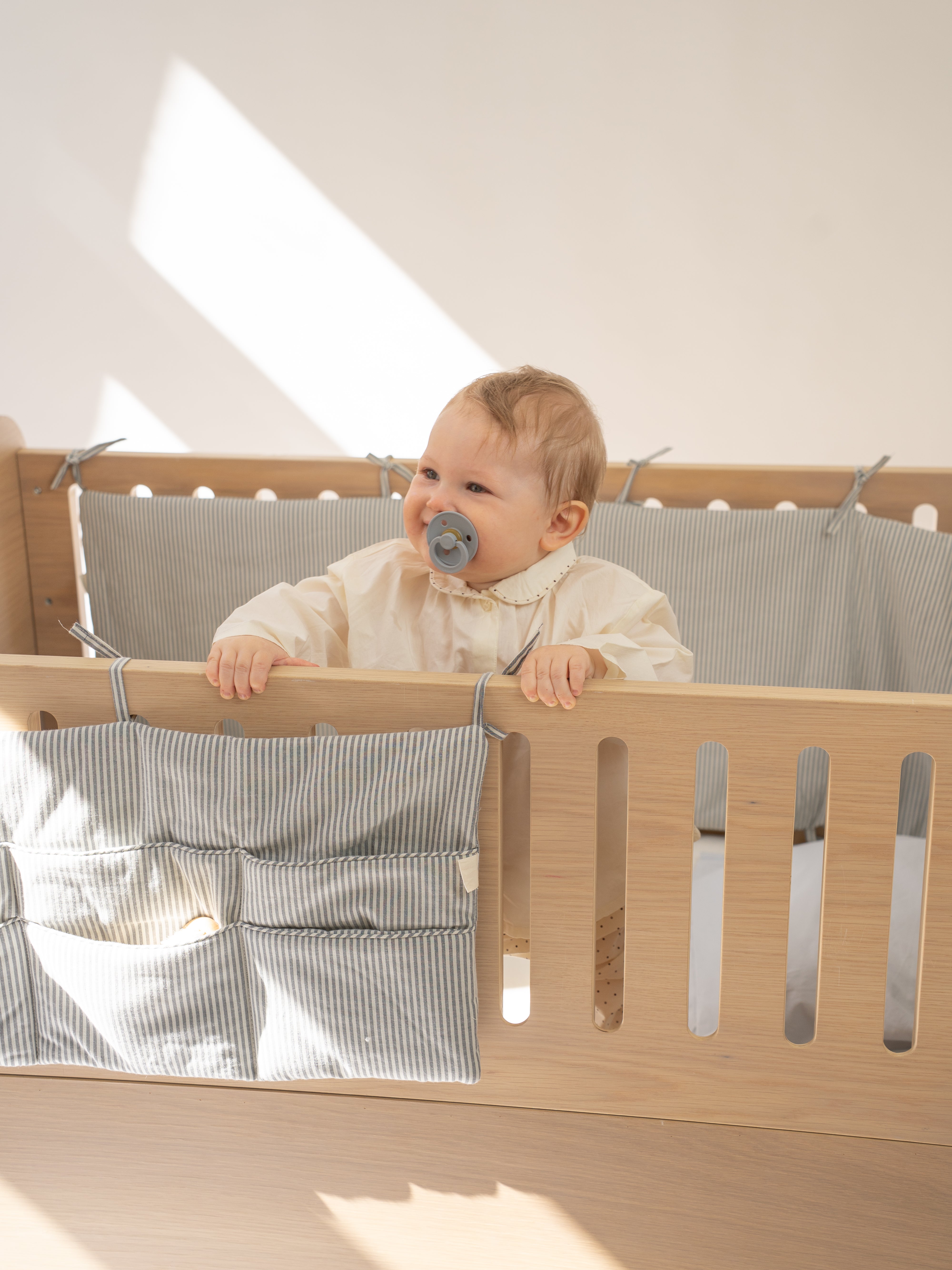 Baby in a crib with a pacifier, smiling in a bright room.