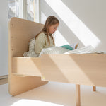 Child reading a book on a wooden bed in a bright room with large windows.