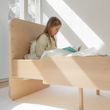Child reading a book on a wooden bed in a bright room with large windows.