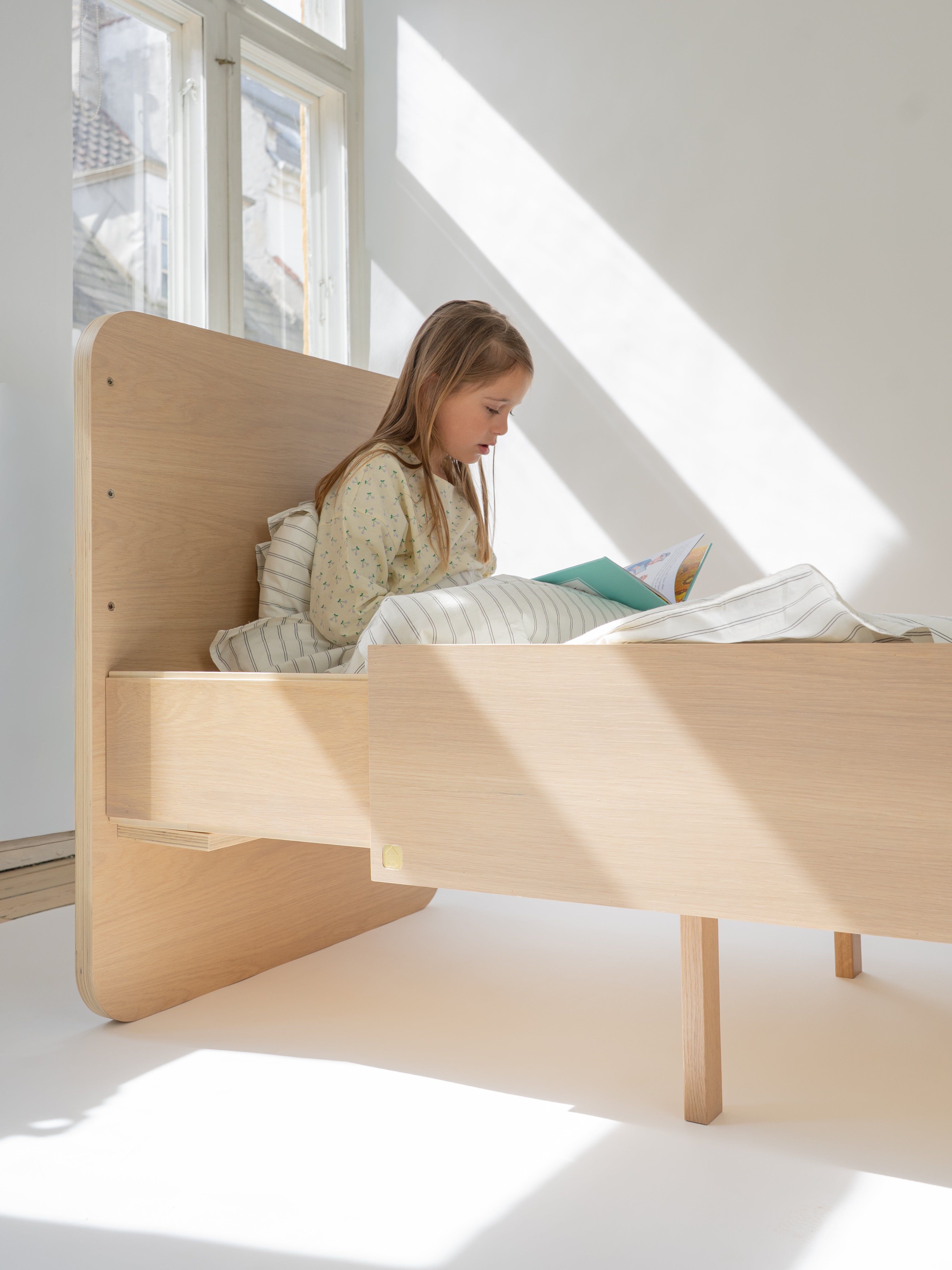 Child reading a book on a wooden bed in a bright room with large windows.