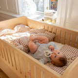 Baby lying in a wooden crib with checkered bedding, holding a teething toy.