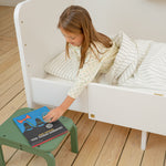 Child sitting on a white bed with a green stool next to them, reading a book.