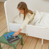 Child sitting on a white bed with a green stool next to them, reading a book.