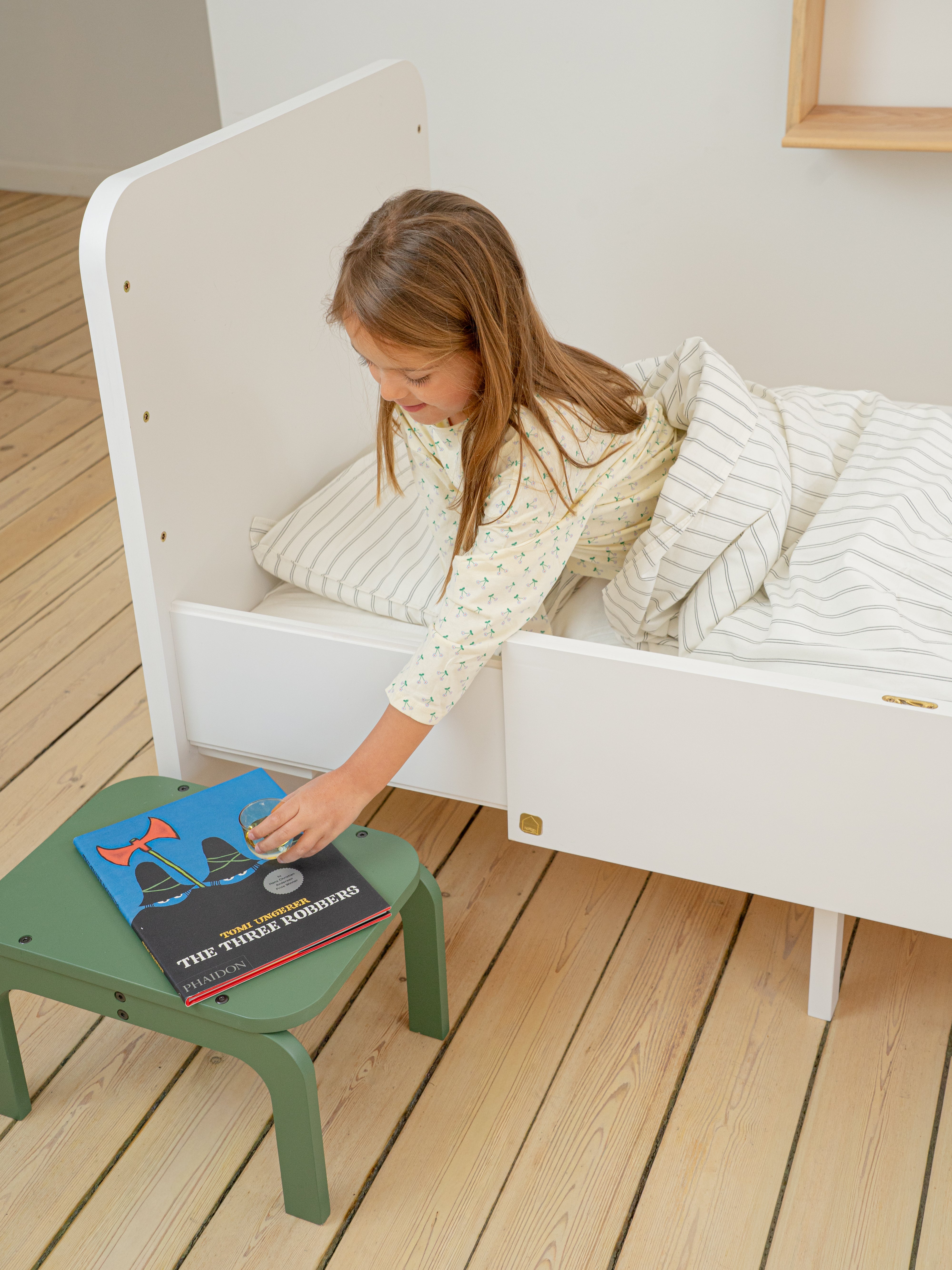 Child sitting on a white bed with a green stool next to them, reading a book.