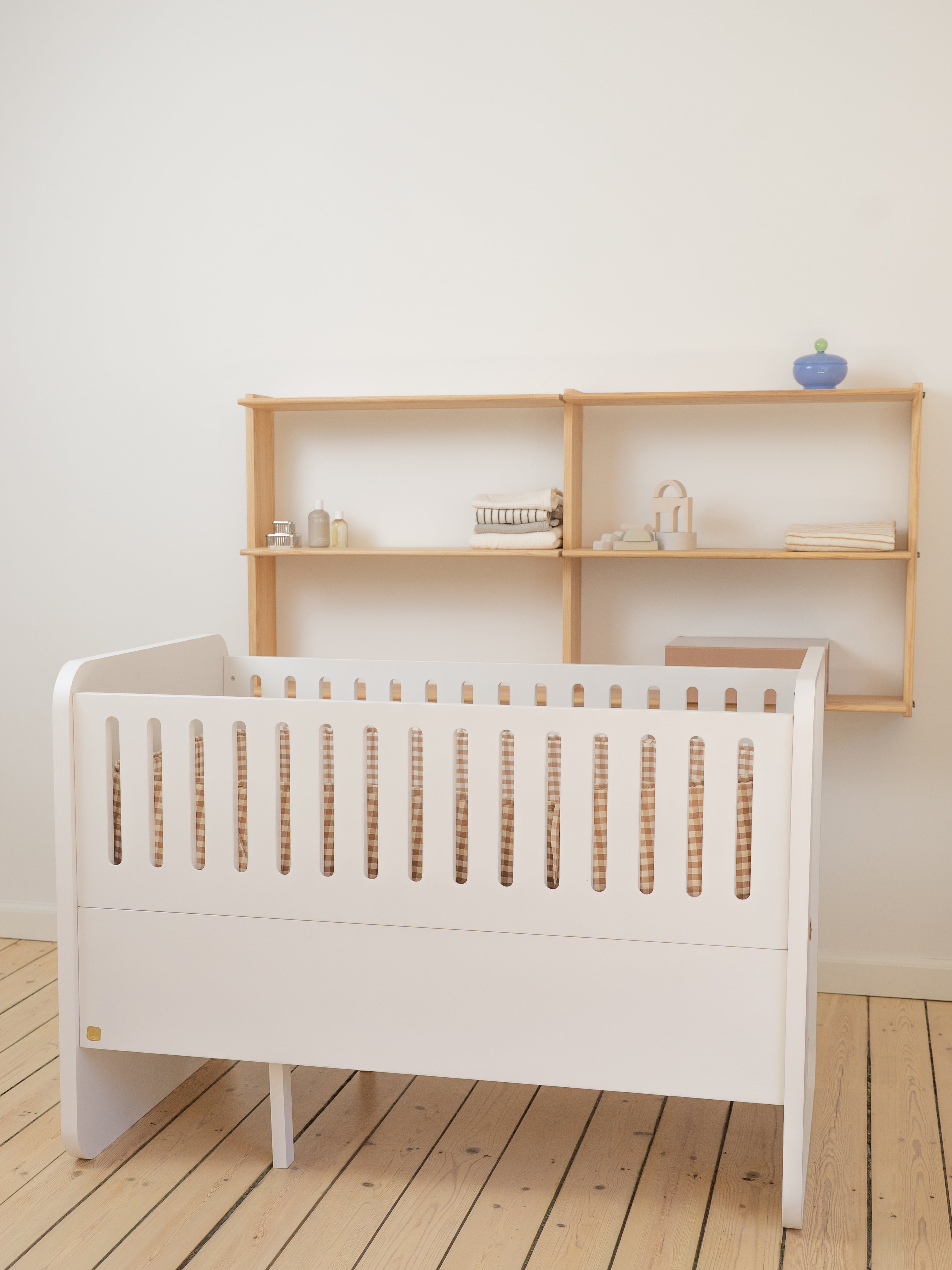 White crib with wooden slats and a wooden shelf unit in a minimalistic room.