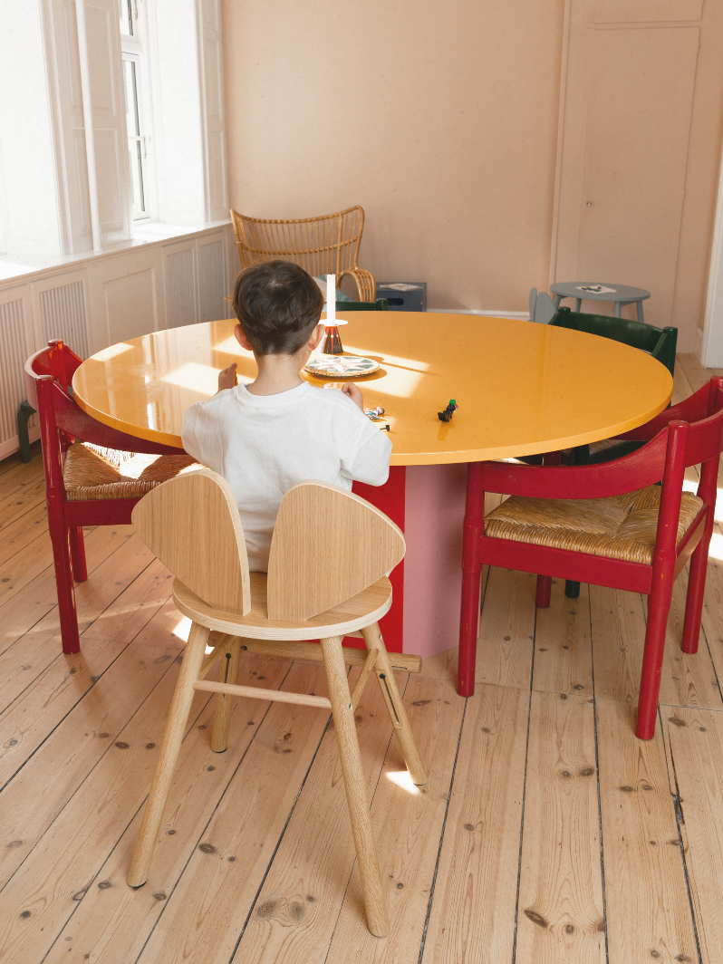 Child sitting at a yellow table with red chairs in a room with wooden flooring.