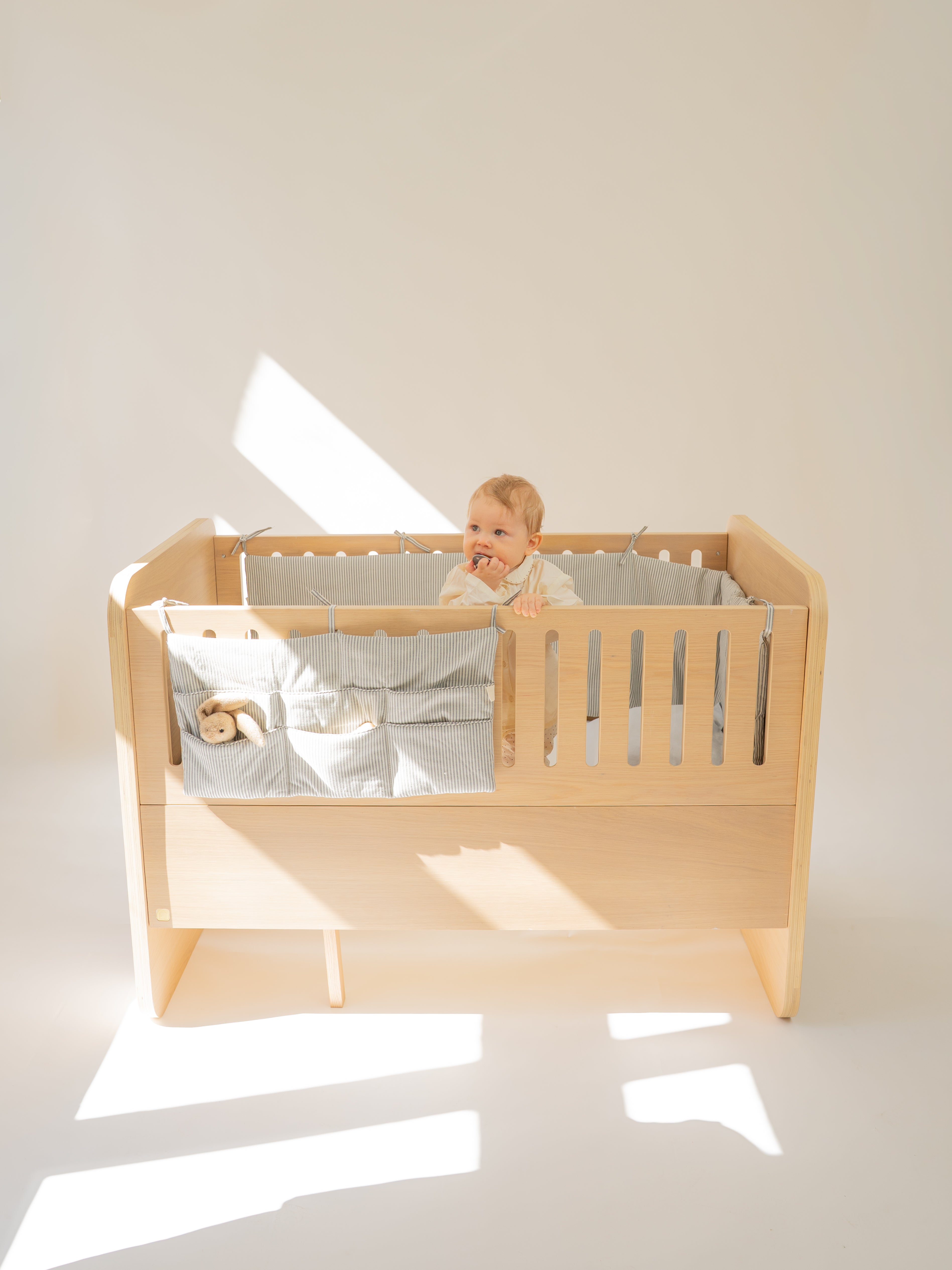 A baby sits inside a light natural wood crib with gray bedding and a matching fabric storage pocket on the side, photographed in a minimalist studio with warm sunlight and shadows.