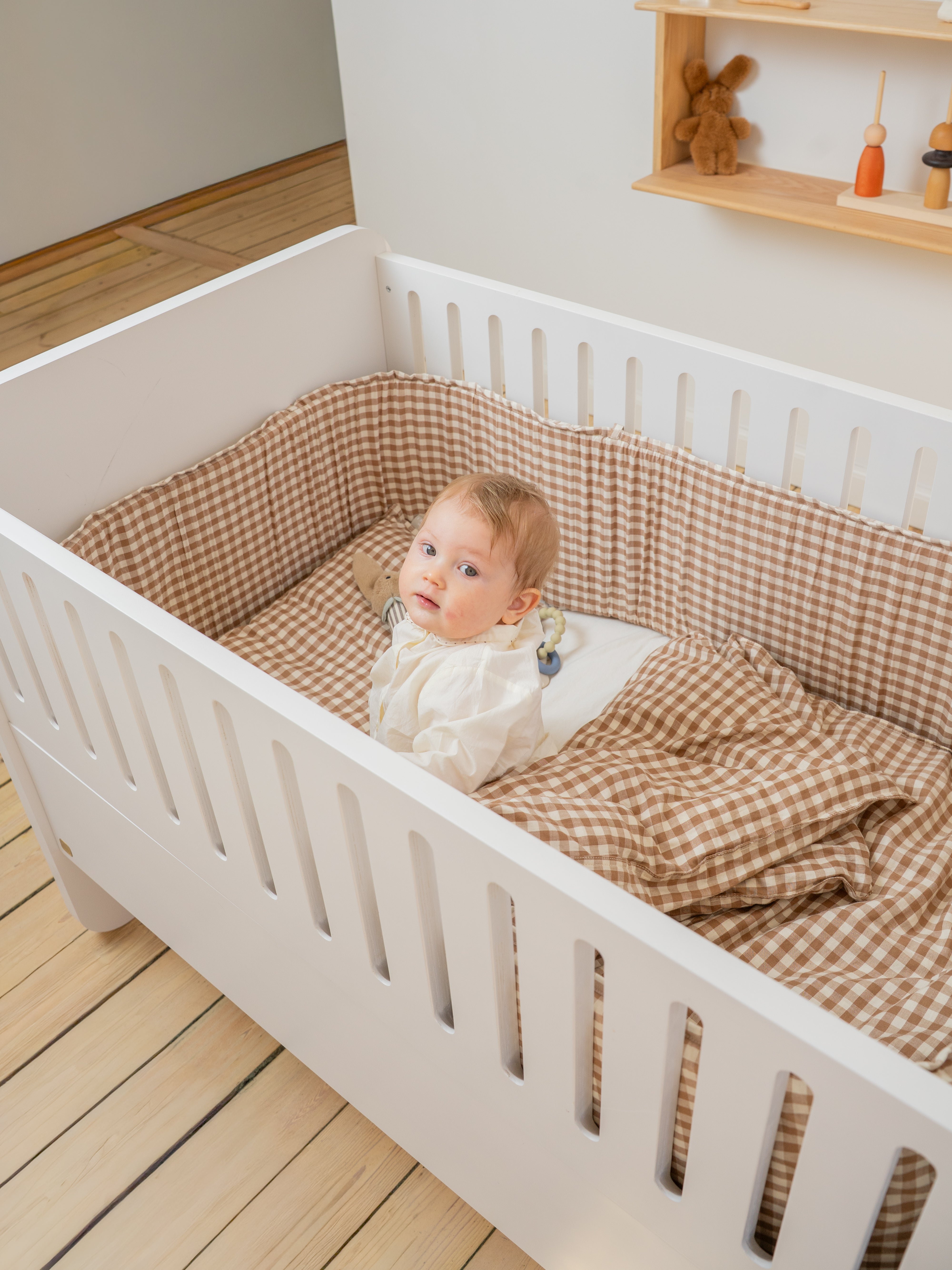 Baby lying in a white crib with brown checkered bedding in a room with wooden floor and shelves.