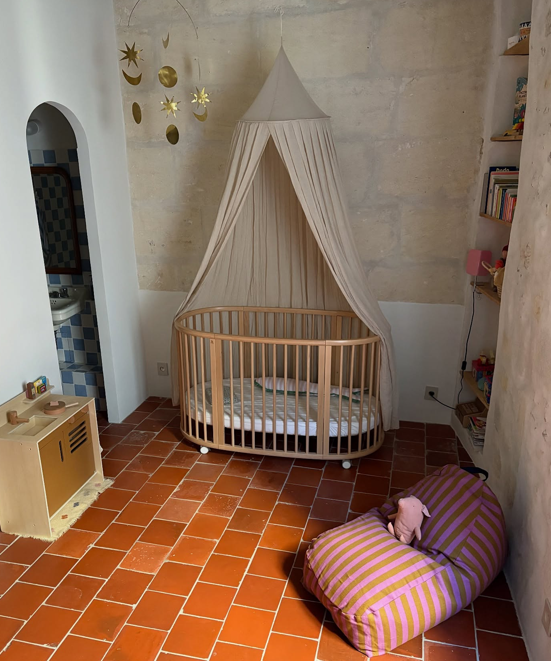 A round wooden baby crib with a beige canopy tent, set in a rustic room with terracotta tiled floors and a striped beanbag chair.