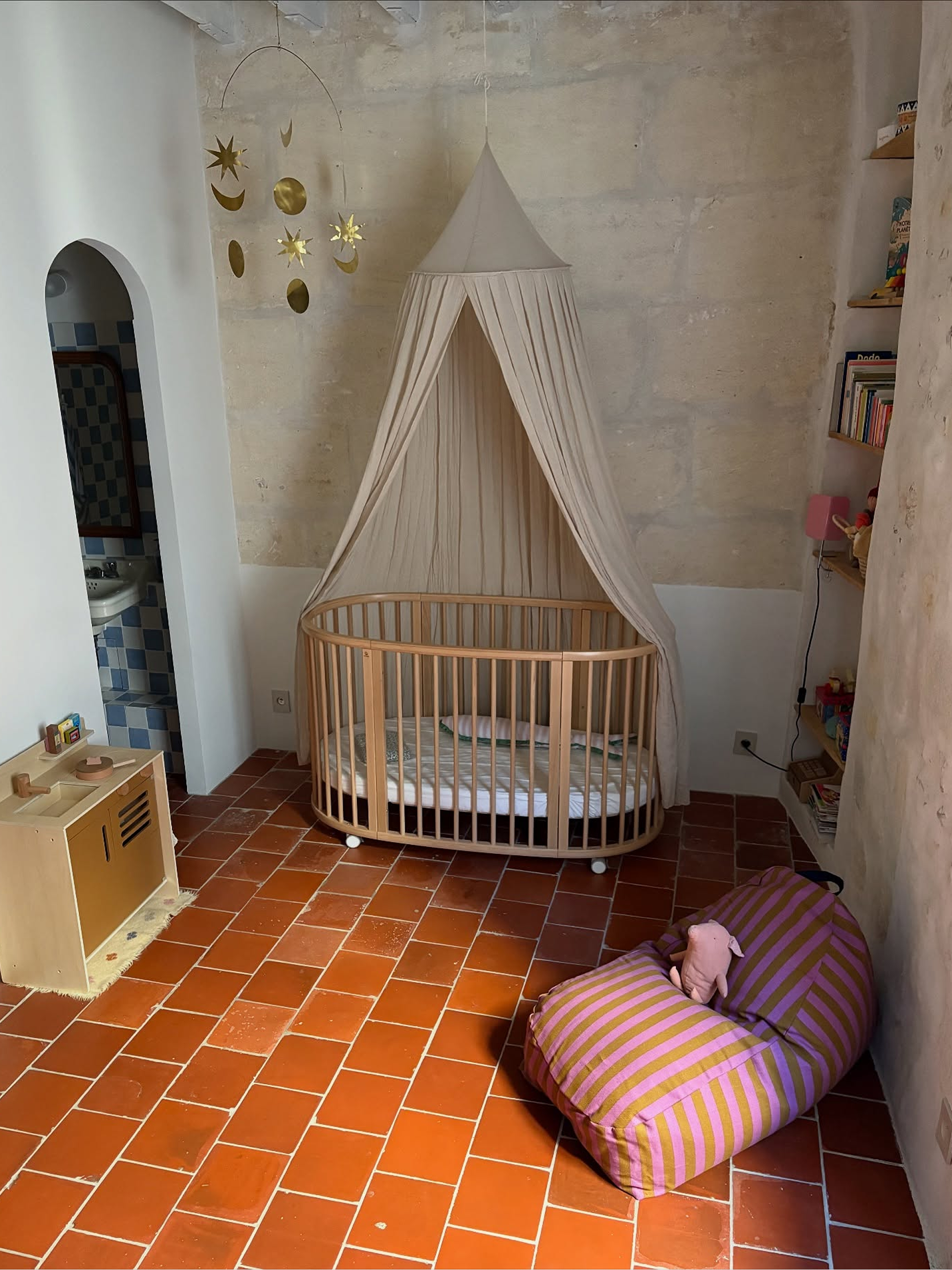 A round wooden baby crib with a beige canopy tent, set in a rustic room with terracotta tiled floors and a striped beanbag chair.
