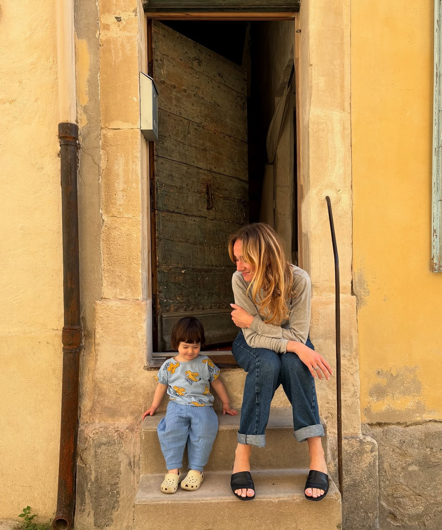 A woman and a young child sit on the stone steps of a rustic yellow building doorway. The woman wears a light gray long-sleeve top, blue jeans, and black slide sandals, while the child wears a light blue patterned top and matching blue pants.
