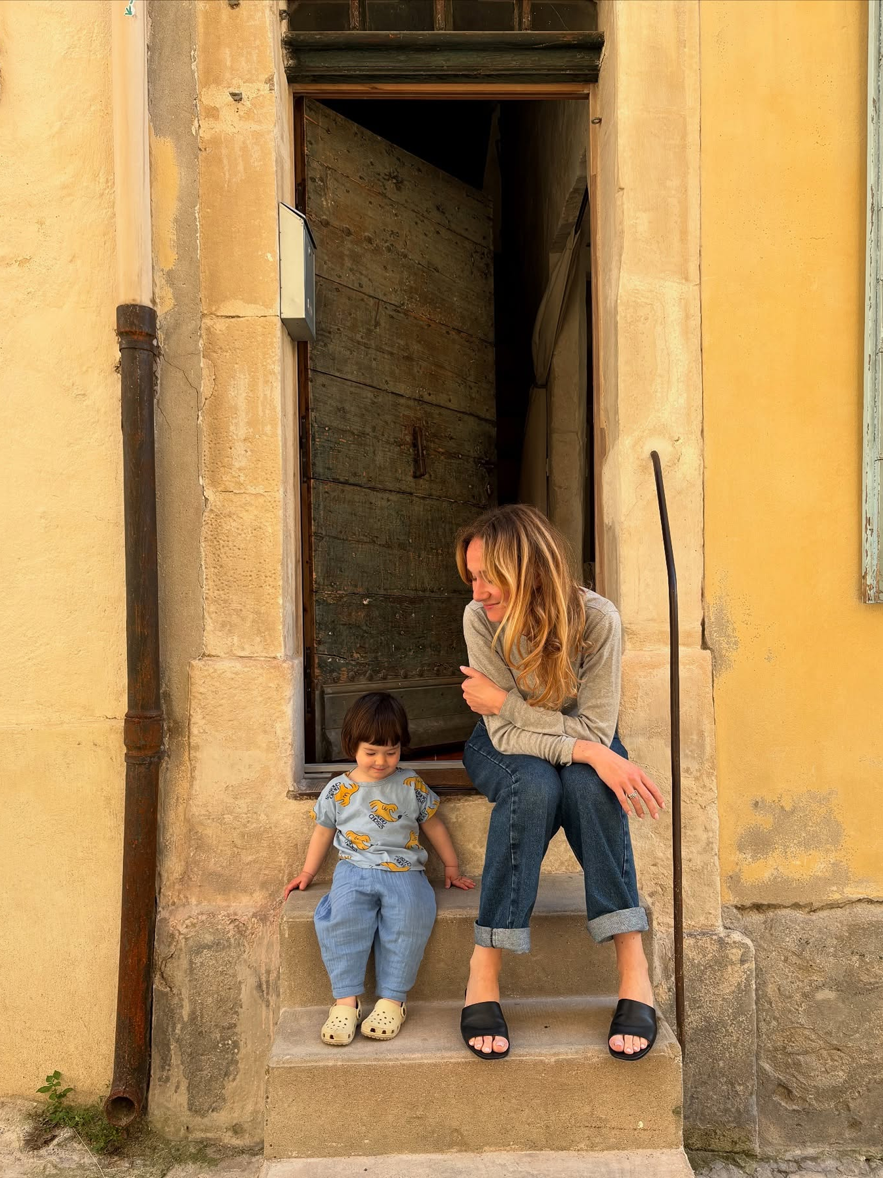 A woman and a young child sit on the stone steps of a rustic yellow building doorway. The woman wears a light gray long-sleeve top, blue jeans, and black slide sandals, while the child wears a light blue patterned top and matching blue pants.