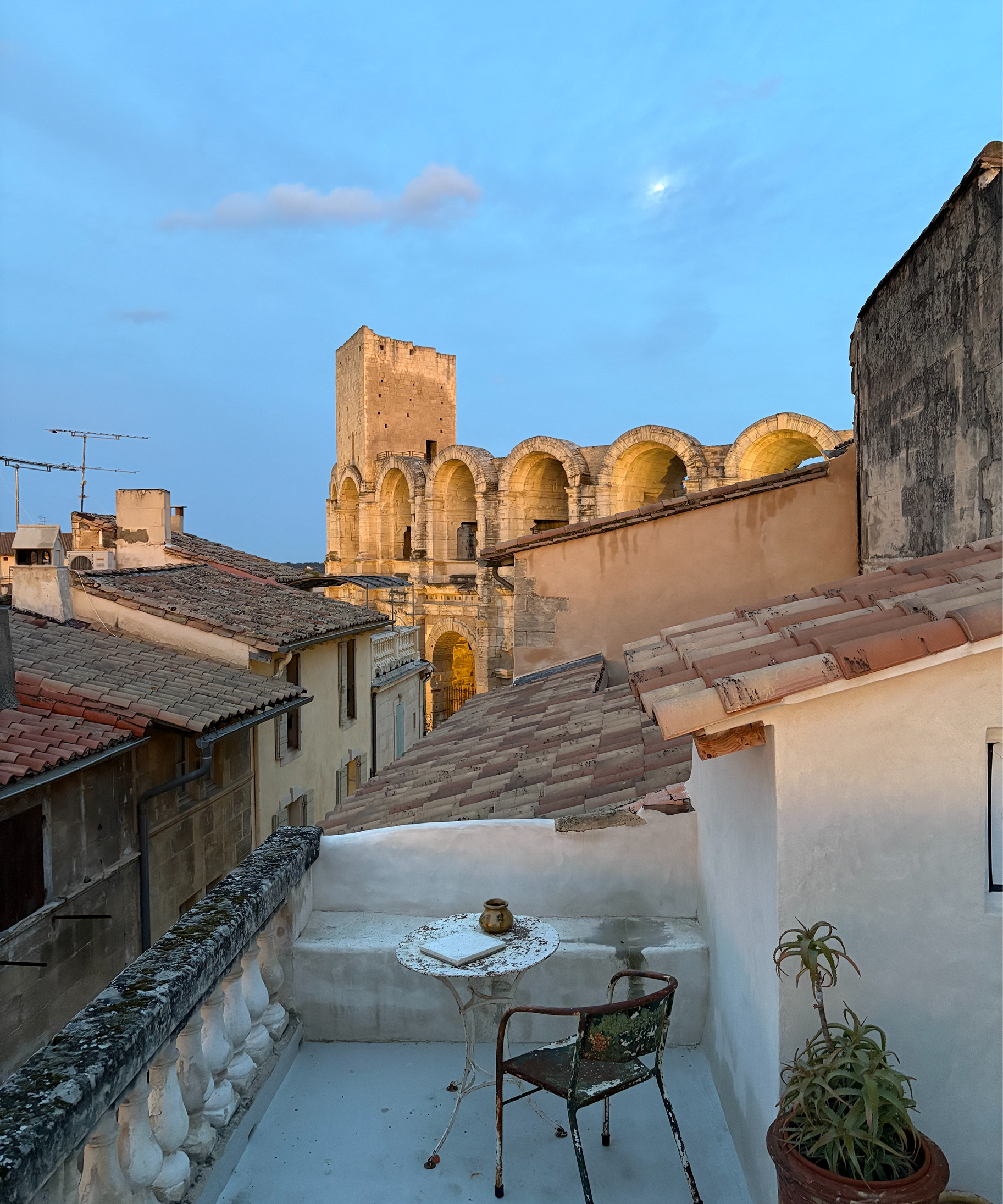 A rooftop terrace with a small round table and chair overlooking a historic stone building with arches under a blue evening sky.