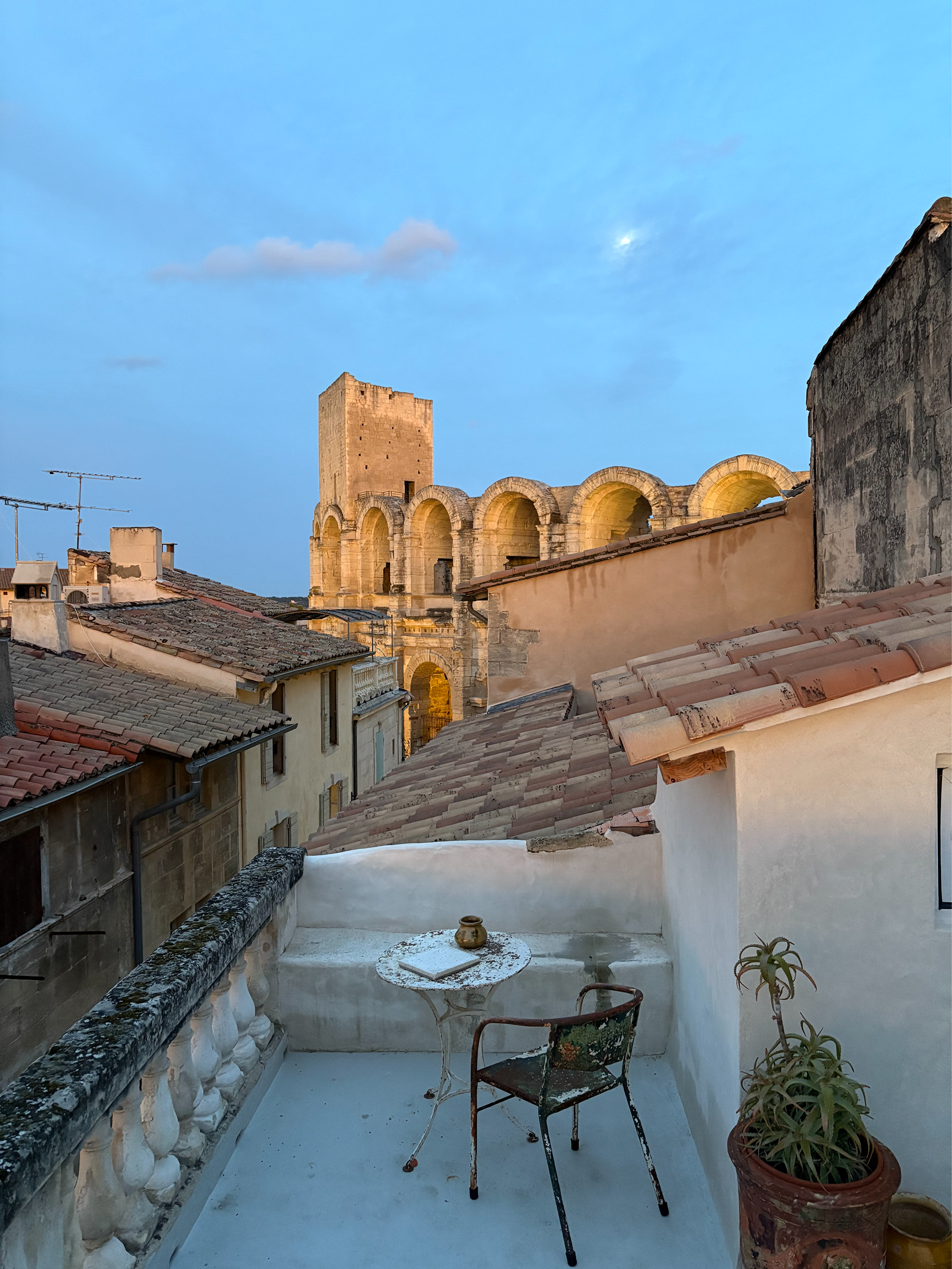 A rooftop terrace with a small round table and chair overlooking a historic stone building with arches under a blue evening sky.