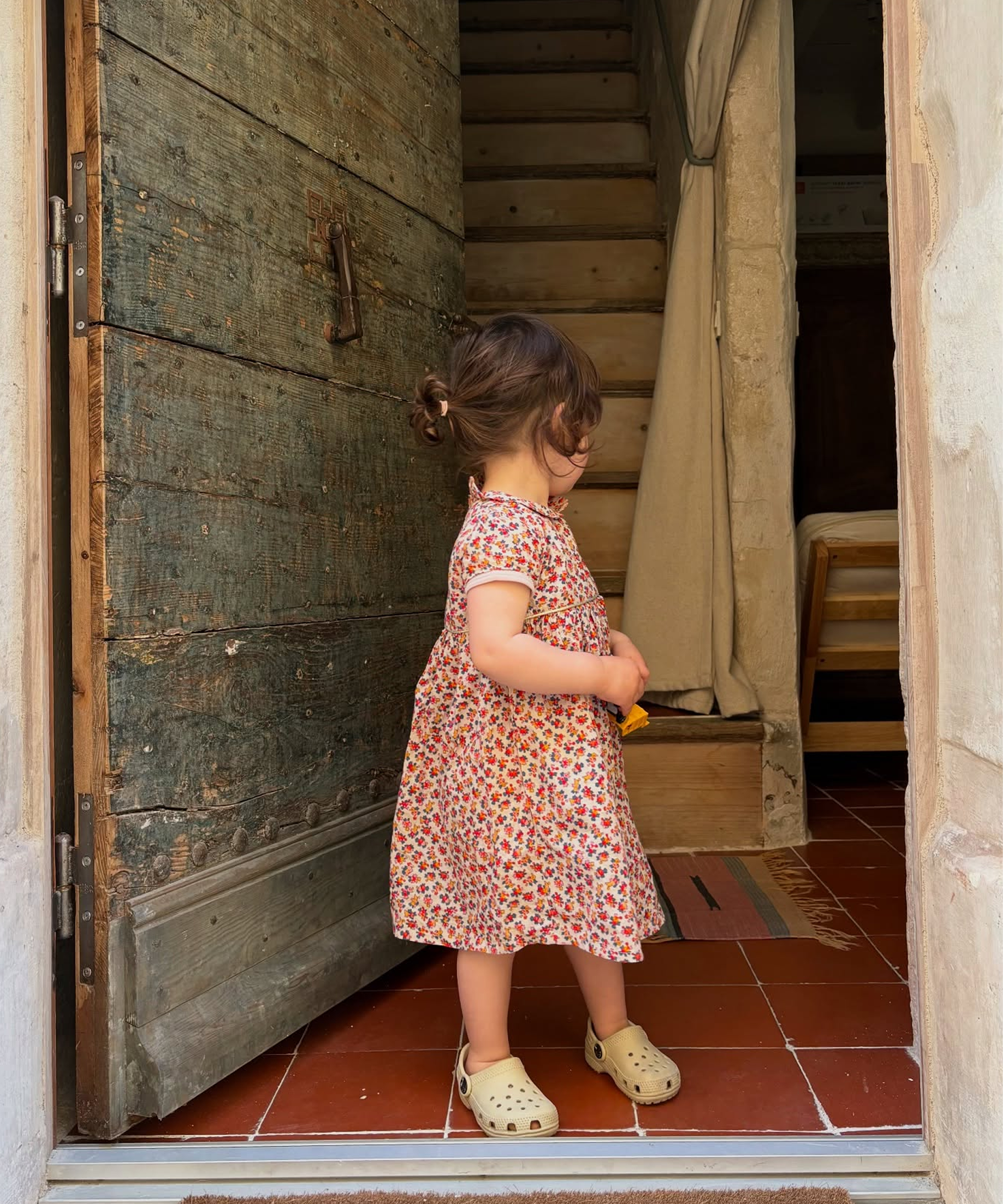 A young child wearing a short-sleeve pink and orange floral print dress and cream clogs stands in a doorway looking down a wooden staircase.