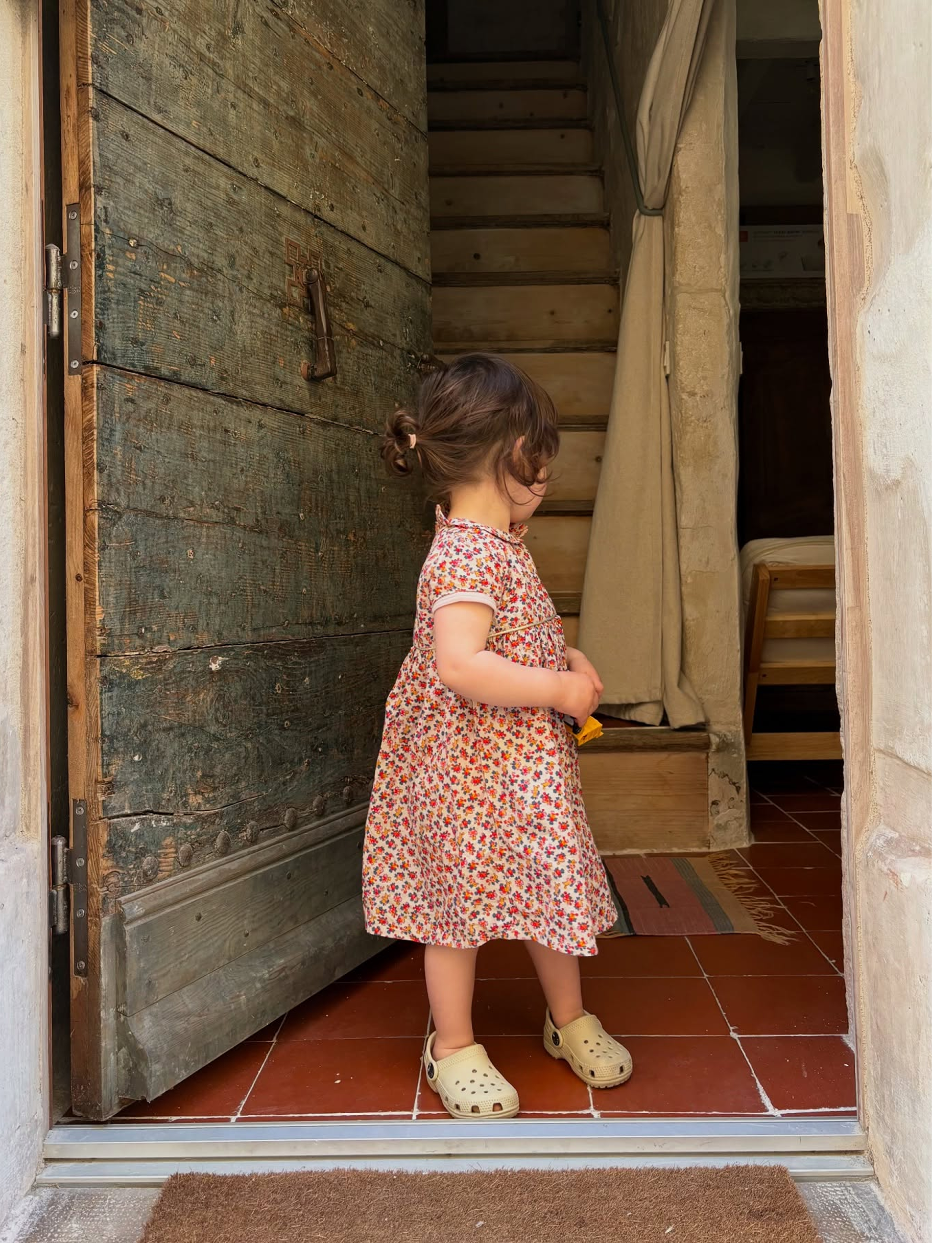 A young child wearing a short-sleeve pink and orange floral print dress and cream clogs stands in a doorway looking down a wooden staircase.