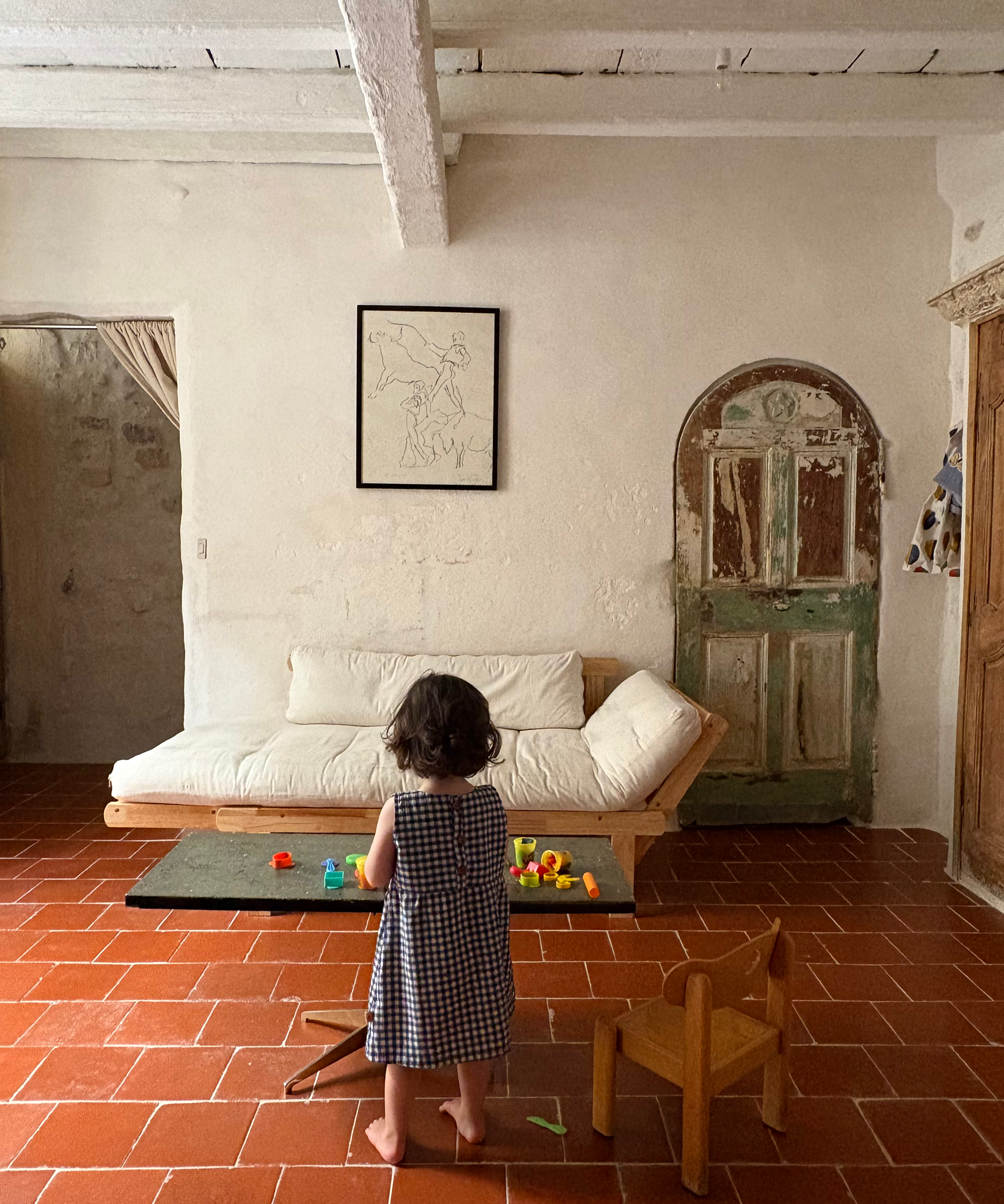 A young child stands barefoot in a rustic room with terracotta tile floors, facing a white daybed and a small table with colorful toys. The child is wearing a black and white checkered dress.