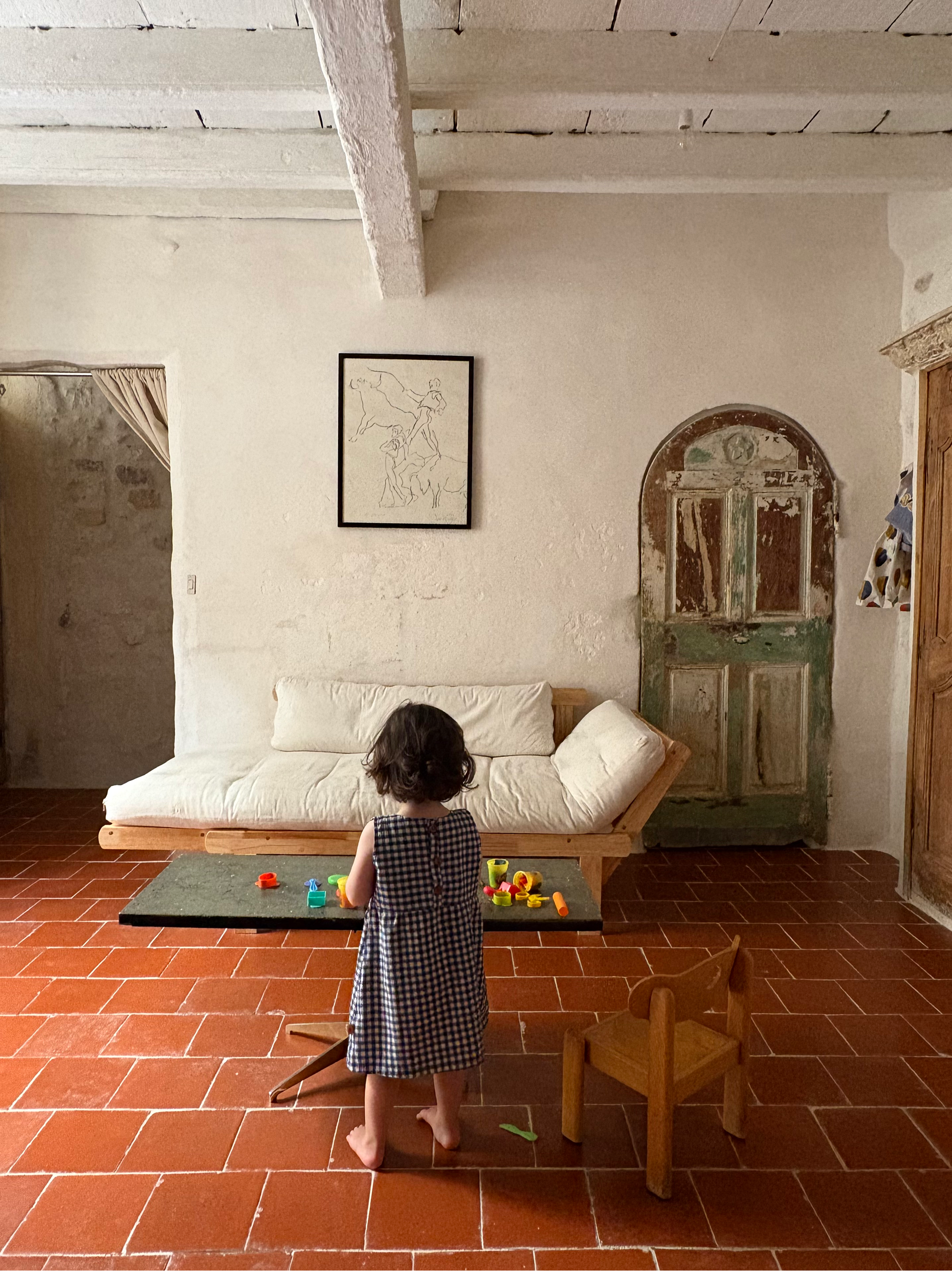A young child stands barefoot in a rustic room with terracotta tile floors, facing a white daybed and a small table with colorful toys. The child is wearing a black and white checkered dress.