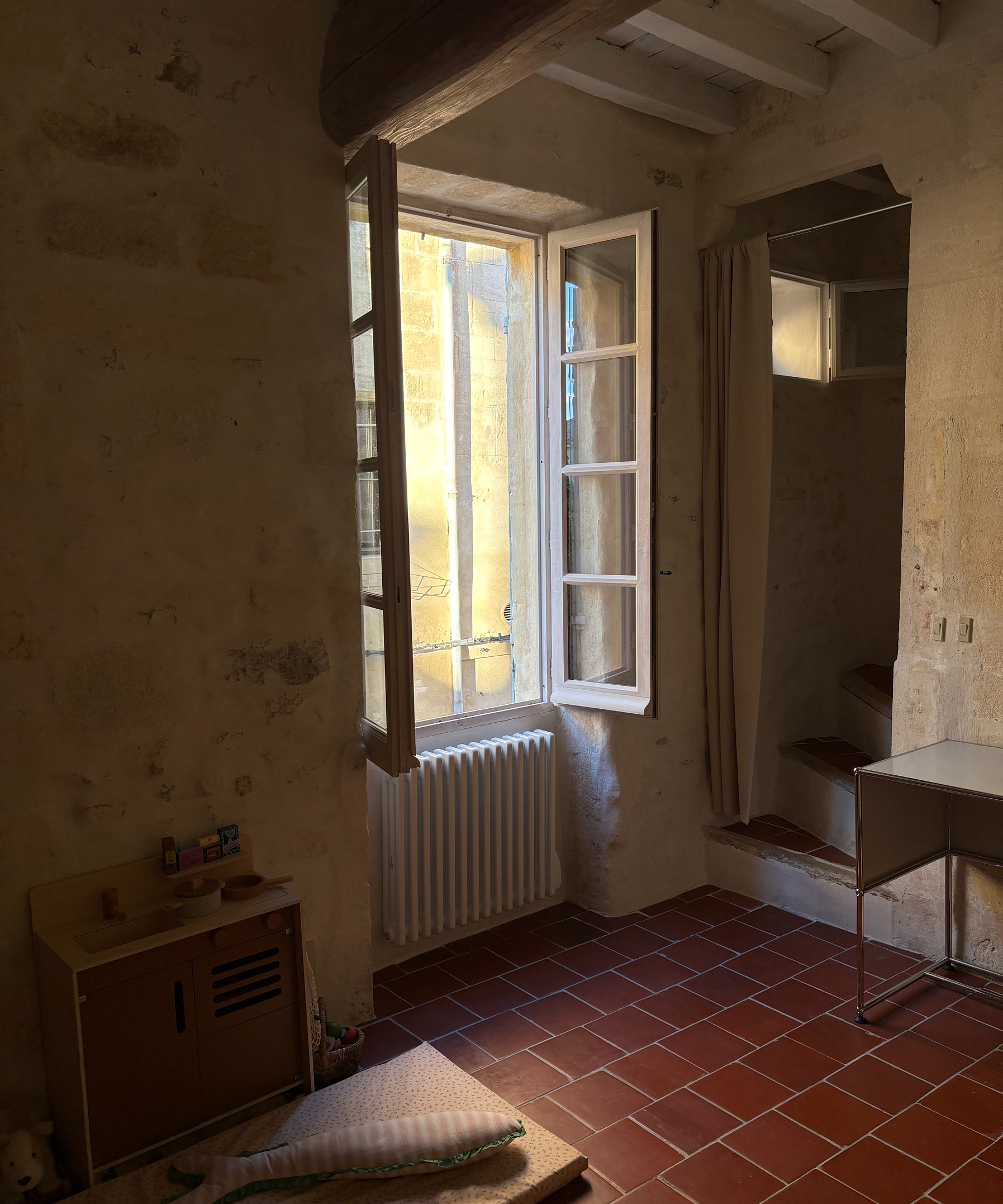 An interior room with rustic stone walls, a red tiled floor, an open white-framed window, a radiator, a small wooden cabinet, and a mirror.