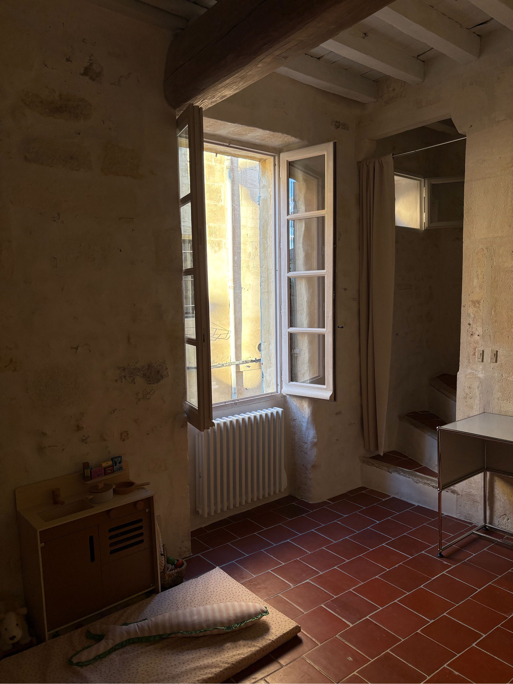 An interior room with rustic stone walls, a red tiled floor, an open white-framed window, a radiator, a small wooden cabinet, and a mirror.