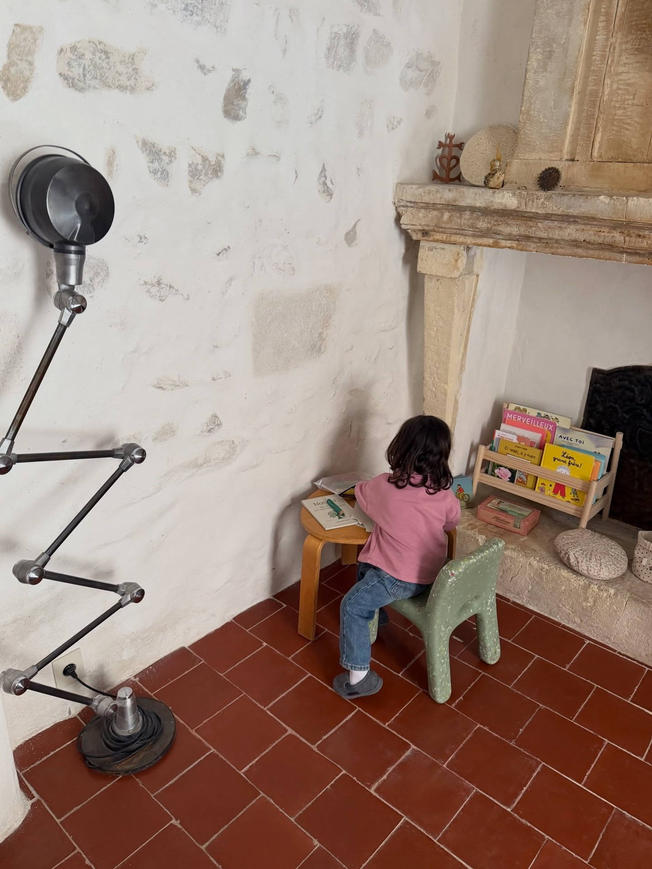 A child sits at a small wooden table on a mint green plastic chair, reading in a rustic room with a stone fireplace, terracotta tile floor, and a black floor lamp on the left.