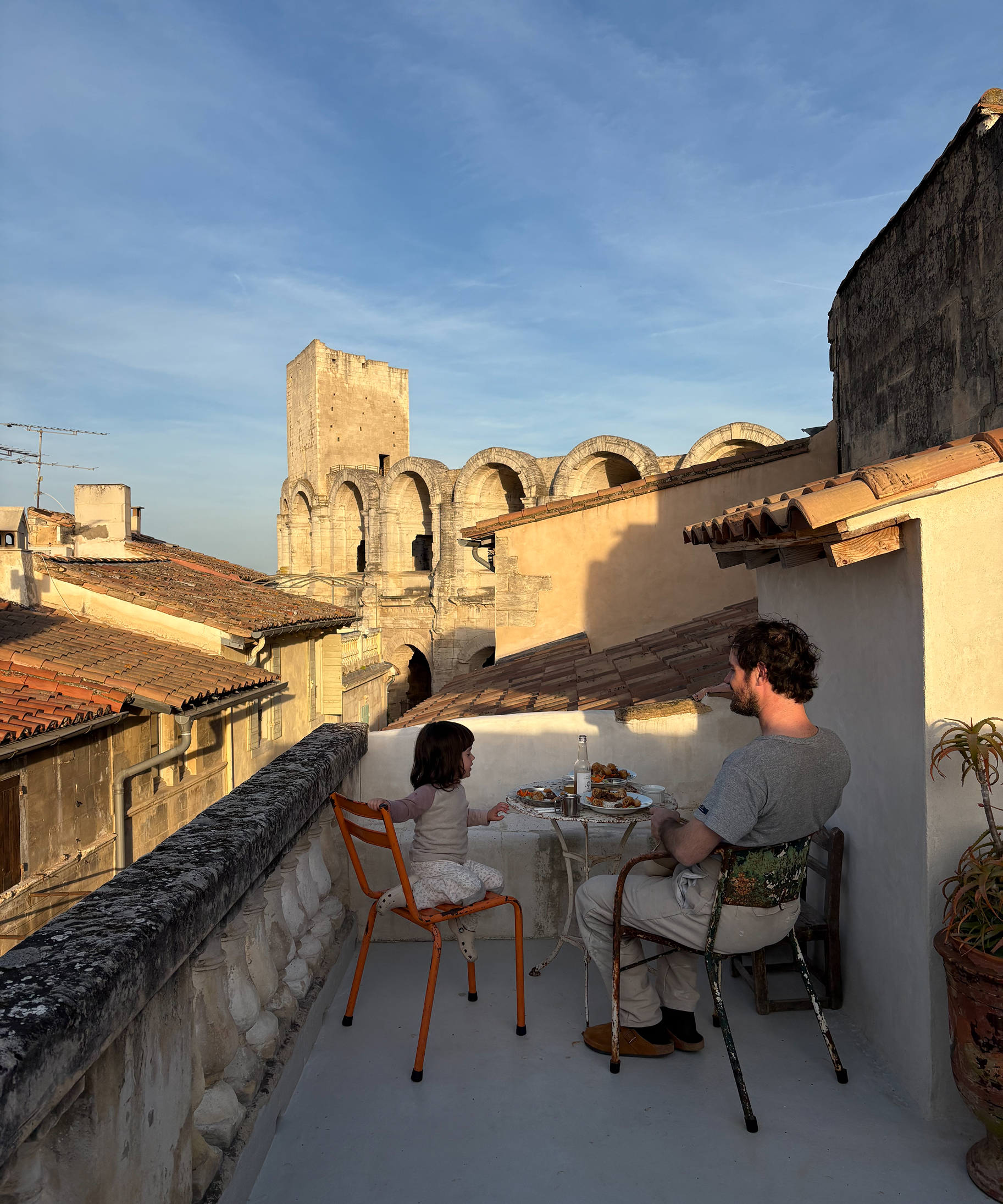 A man and a young girl sit at a small table on a rooftop terrace, eating outdoors with a view of historic stone buildings and a blue sky. The man wears a gray t-shirt and light pants, and the child wears a light pink outfit.