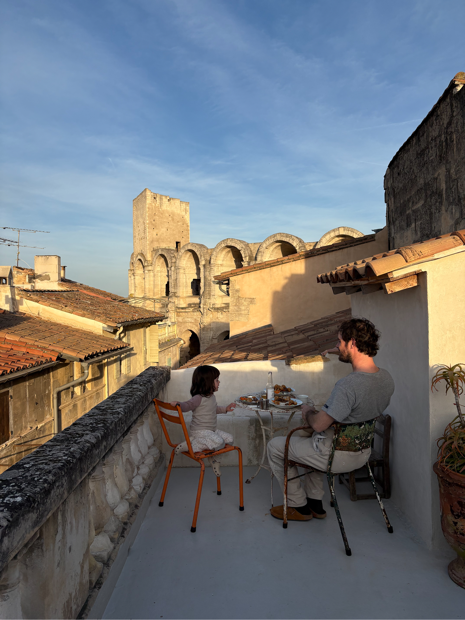 A man and a young girl sit at a small table on a rooftop terrace, eating outdoors with a view of historic stone buildings and a blue sky. The man wears a gray t-shirt and light pants, and the child wears a light pink outfit.