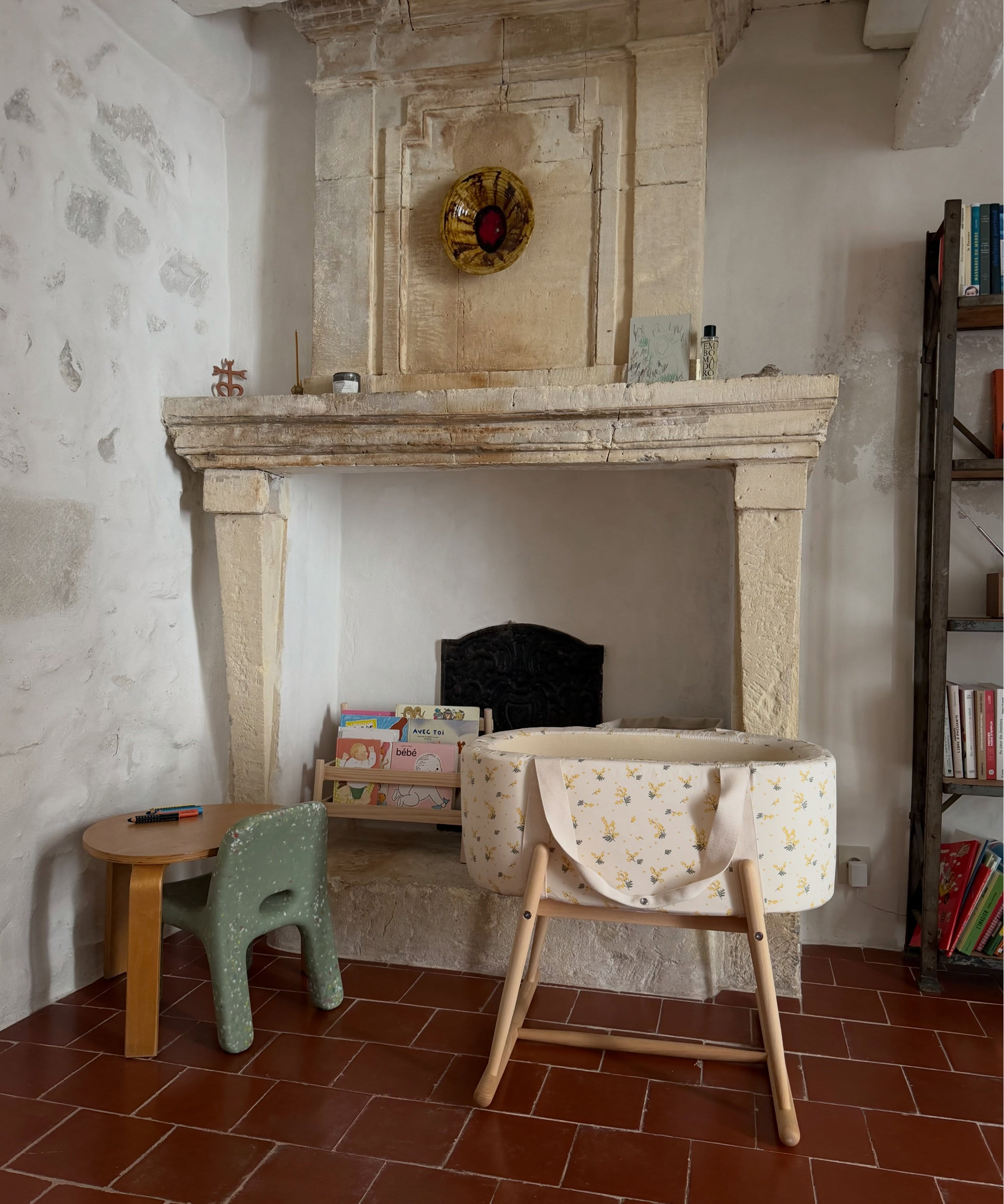 A baby bassinet with a cream floral print and wooden legs sits in front of a large stone fireplace in a rustic room with terracotta tiled floors.