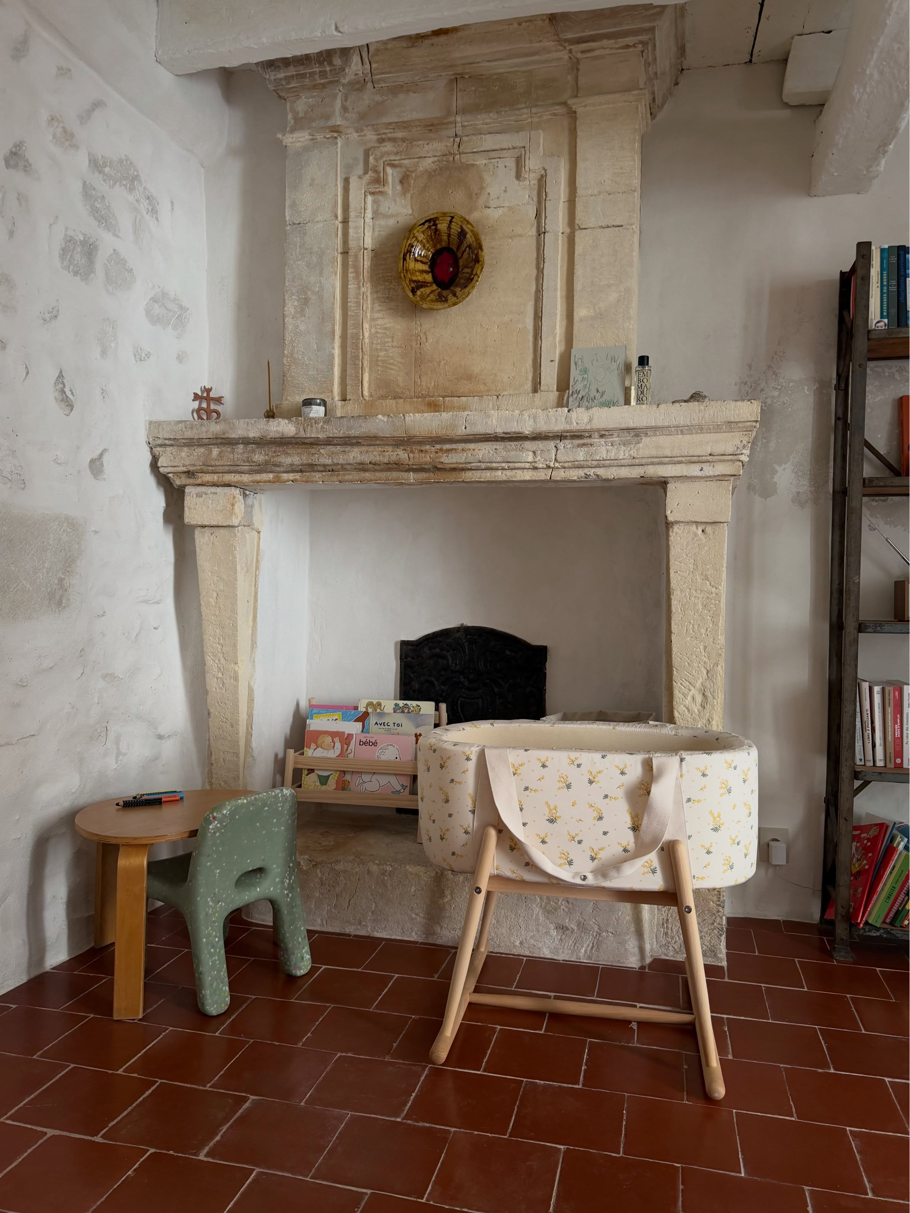 A baby bassinet with a cream floral print and wooden legs sits in front of a large stone fireplace in a rustic room with terracotta tiled floors.