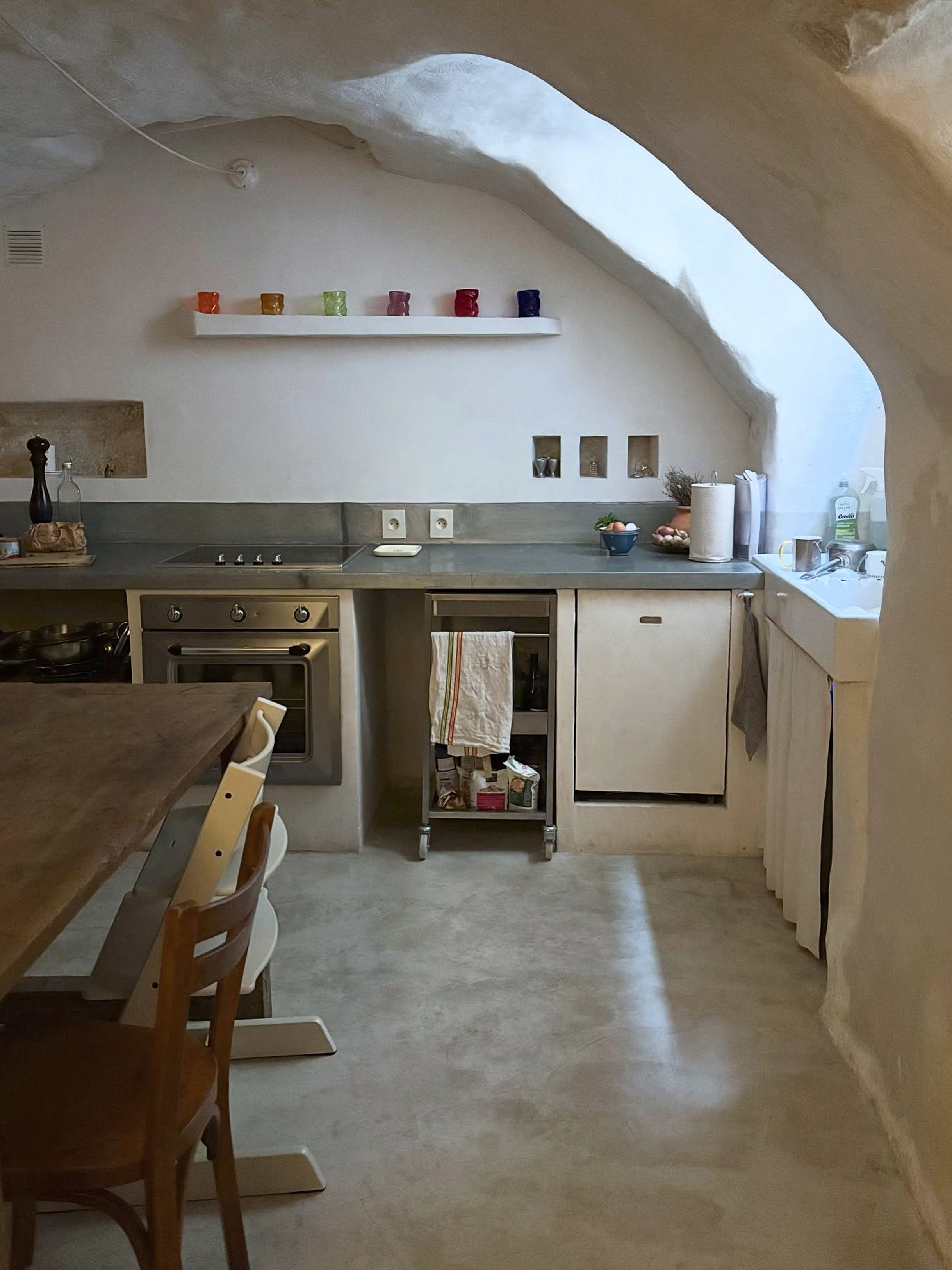 A rustic kitchen interior with a stone arch ceiling, white walls, a built-in oven, countertop fixtures, and a wooden dining table with chairs in the foreground.