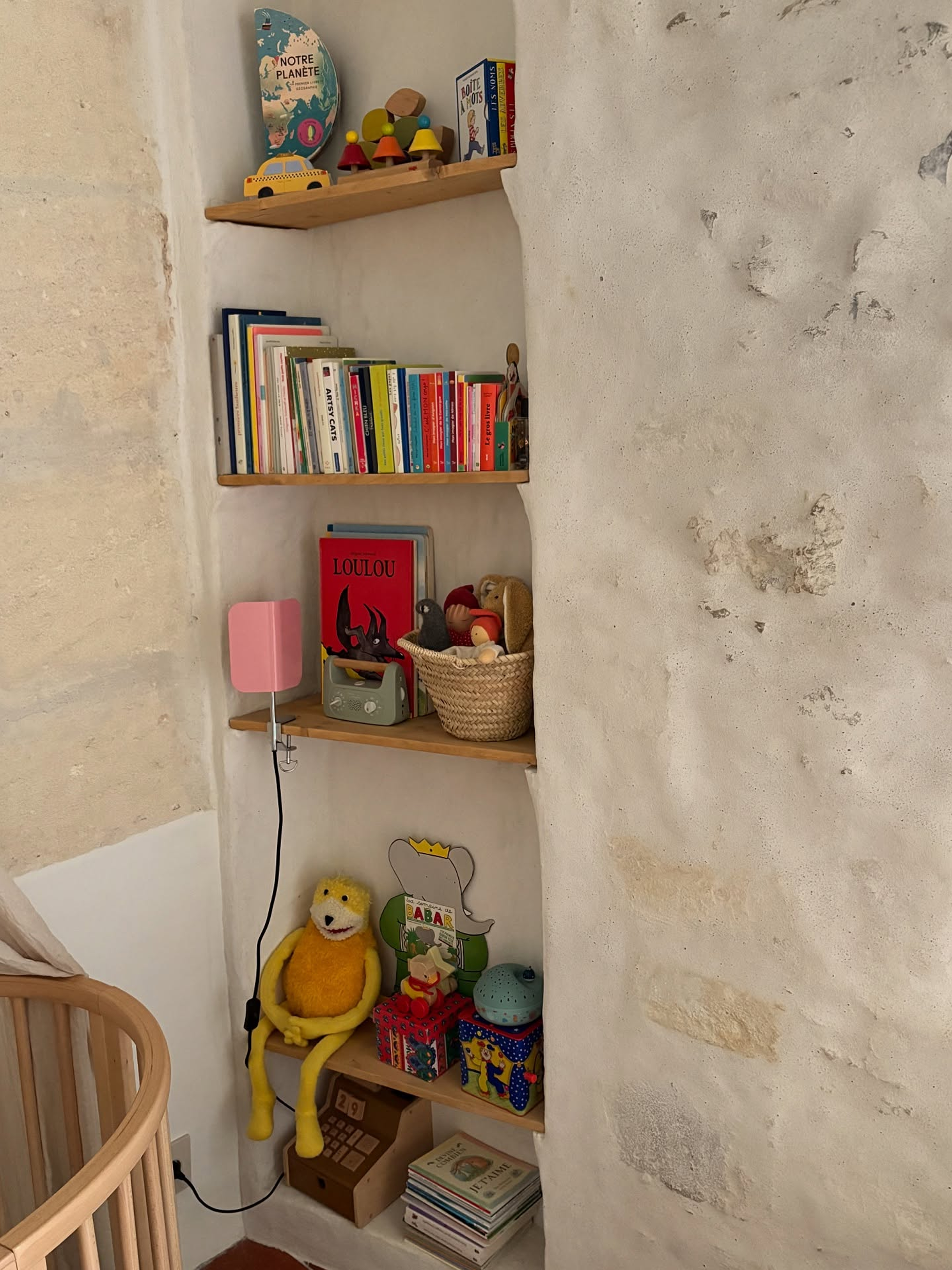 A set of four light wood floating shelves mounted on a textured white plaster wall, styled with children's books, toys, a pink speaker, and a yellow plush toy in a cozy nursery or playroom corner.