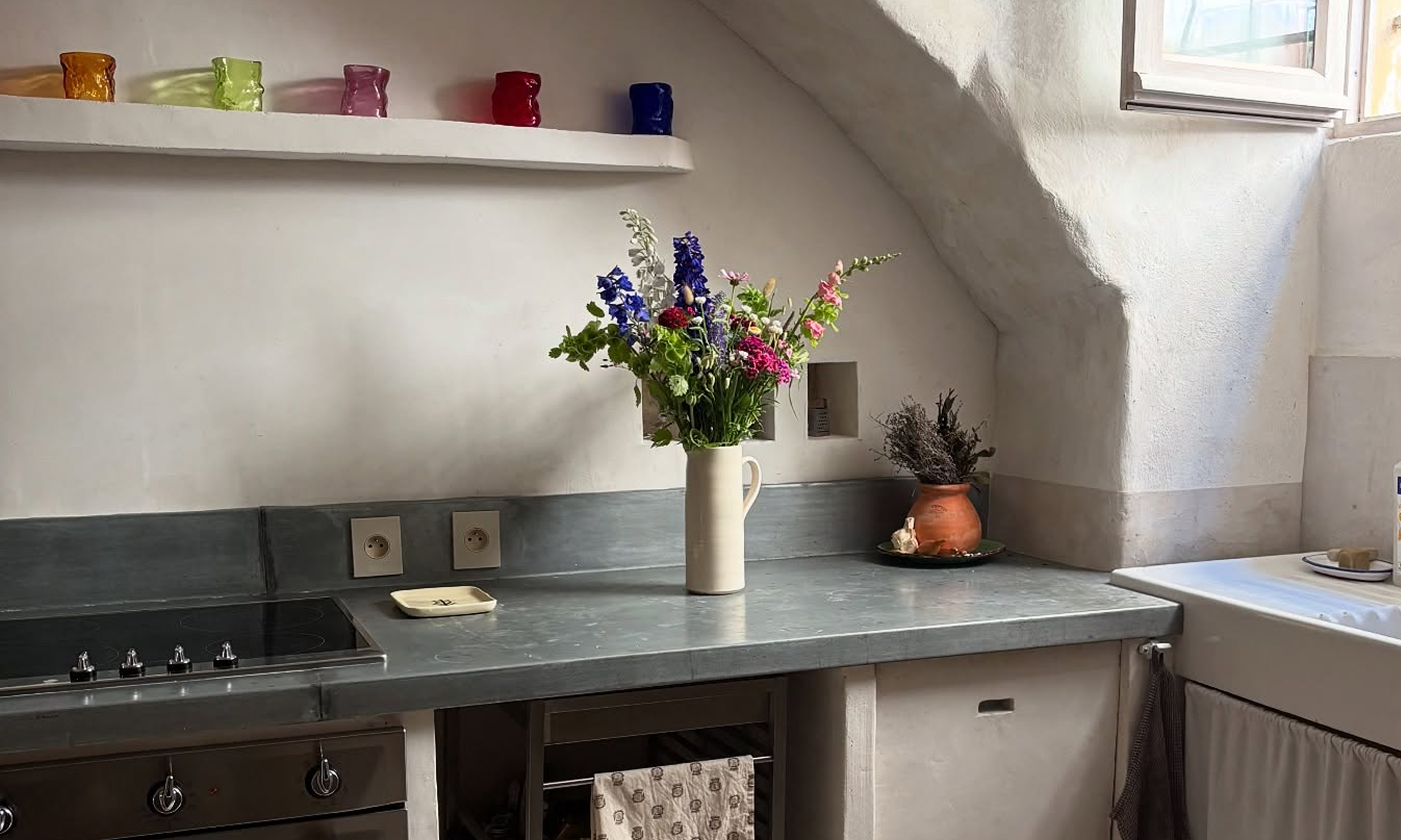 A rustic kitchen scene with a gray countertop, cream-colored cabinets, a white shelf with colorful glass cups, and a vase of flowers in the center.
