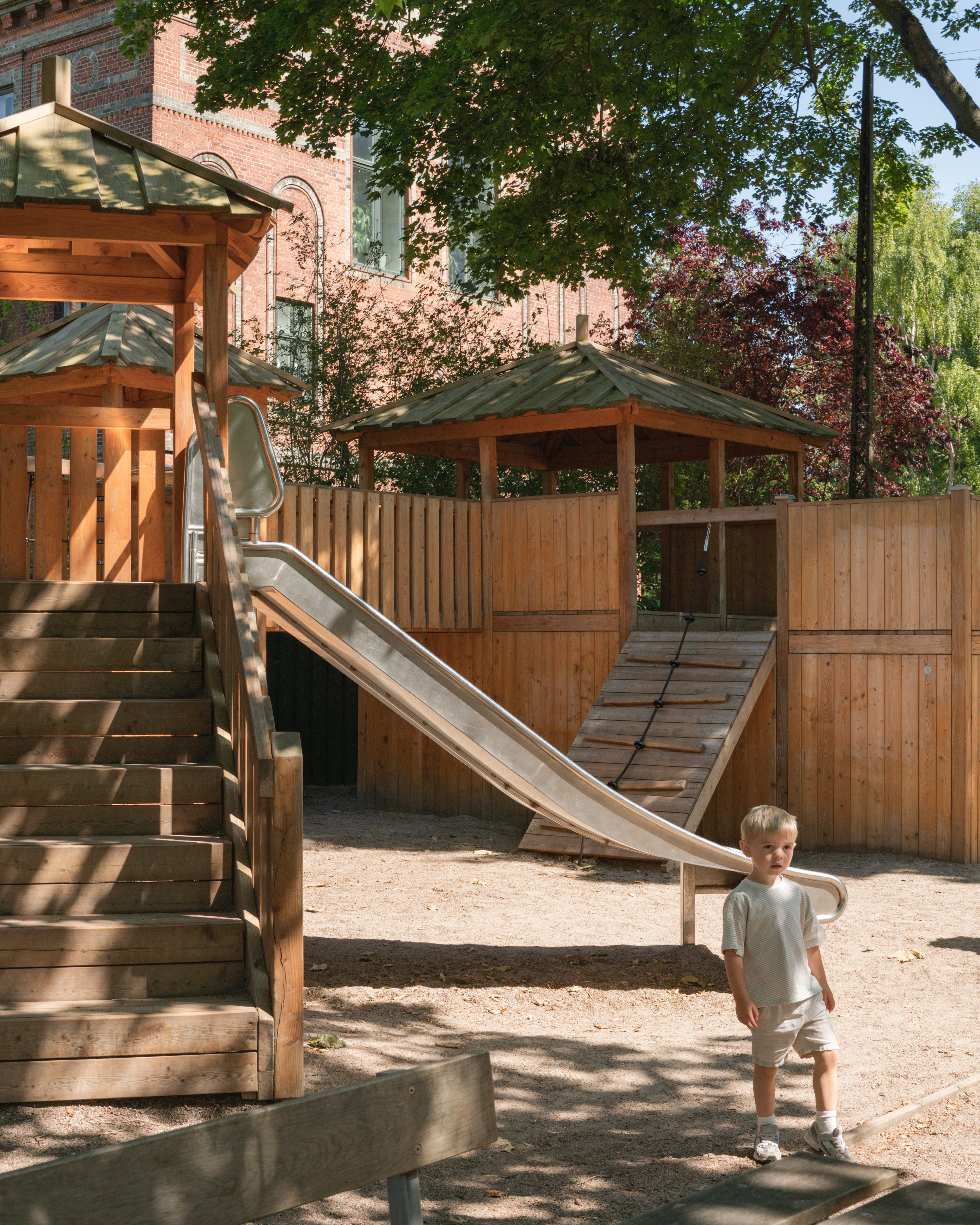 Child standing near a wooden playground structure with slides and steps.