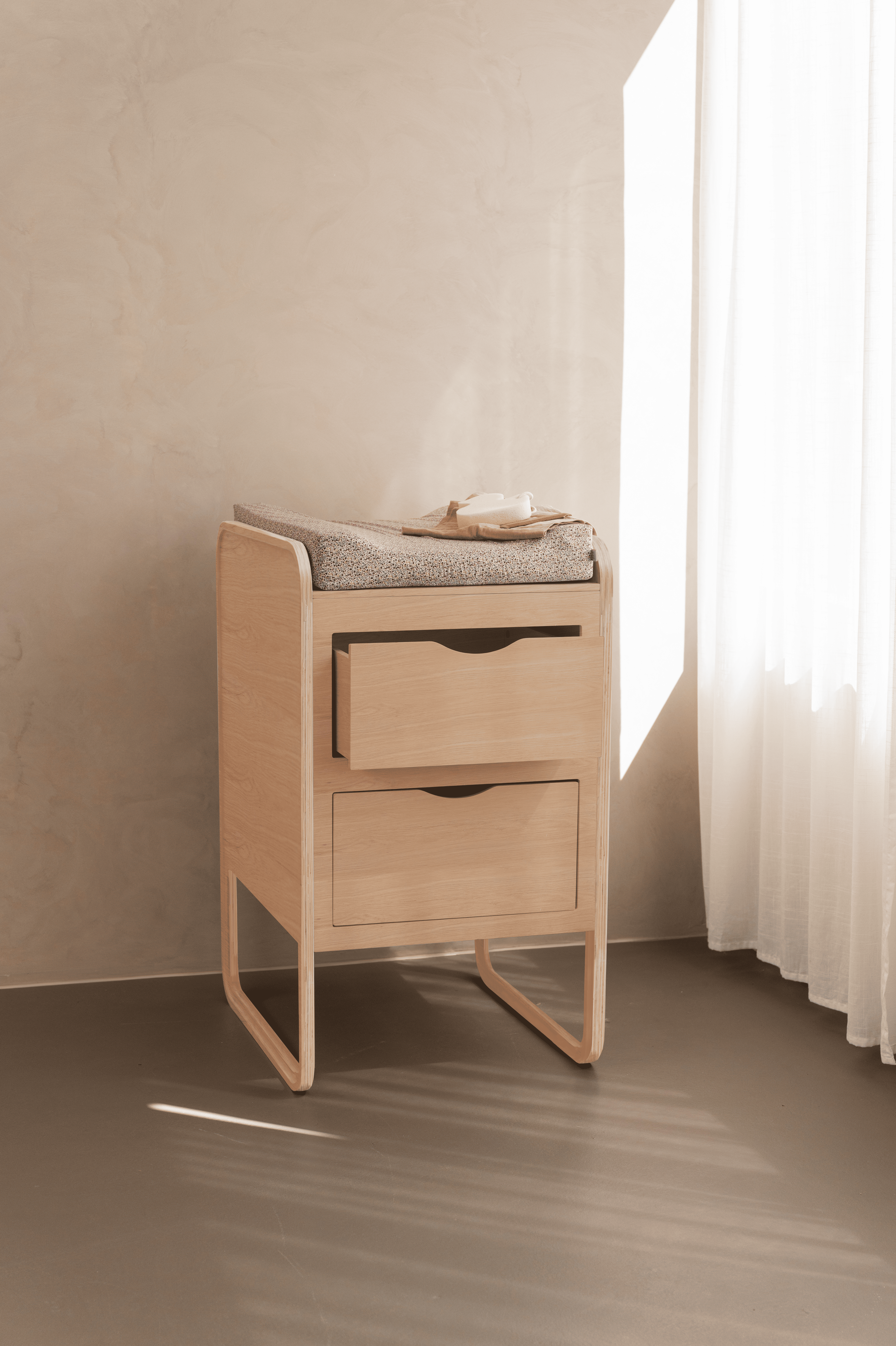 A light natural wood changing table with two drawers and a gray textured pad on top, styled in a minimalist room with warm beige walls, a window, and white sheer curtains.
