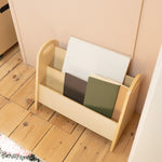 A light natural wood magazine rack with three compartments, holding notebooks, placed on a light wood floor against a pale pink wall.
