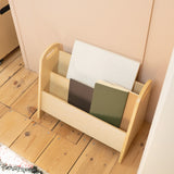A light natural wood magazine rack with three compartments, holding notebooks, placed on a light wood floor against a pale pink wall.
