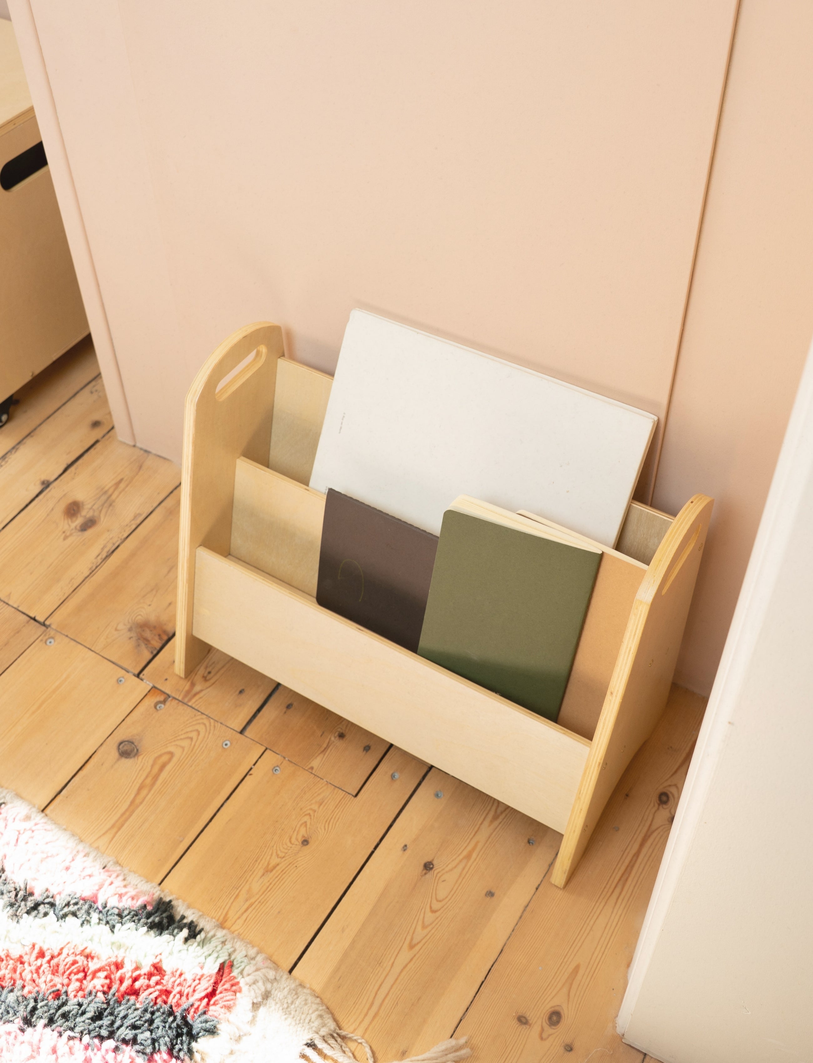 A light natural wood magazine rack with three compartments, holding notebooks, placed on a light wood floor against a pale pink wall.