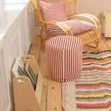 A cozy room corner featuring a natural wicker rocking chair with red and white striped cushions, a matching cylindrical ottoman, a colorful striped rug, and a light wood magazine rack on a wooden floor.