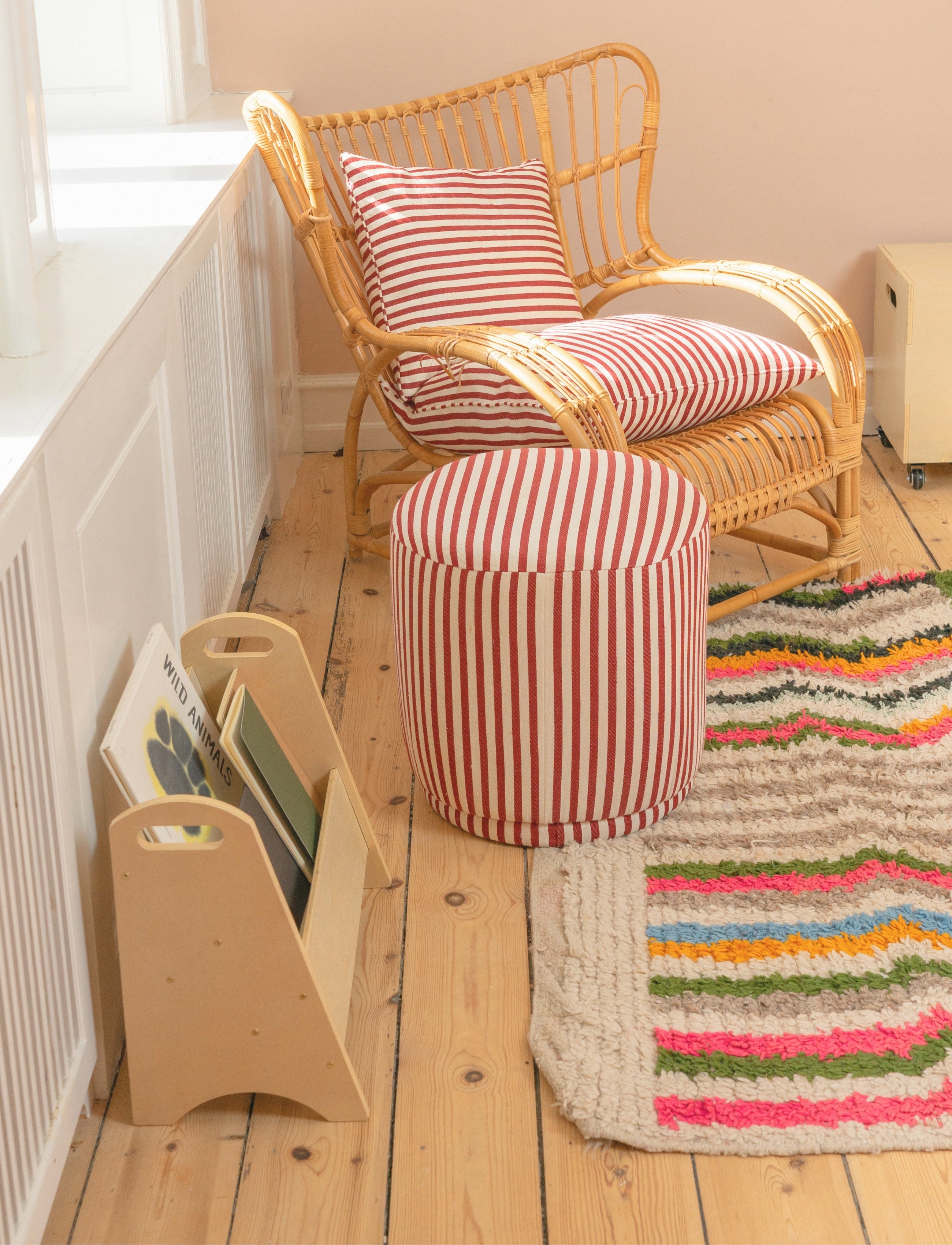 A cozy room corner featuring a natural wicker rocking chair with red and white striped cushions, a matching cylindrical ottoman, a colorful striped rug, and a light wood magazine rack on a wooden floor.