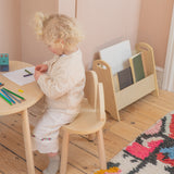 A toddler with curly blonde hair sits at a small round wooden table, drawing on paper. The child wears a cream knit cardigan and cream trousers, sitting on a matching light wood chair.