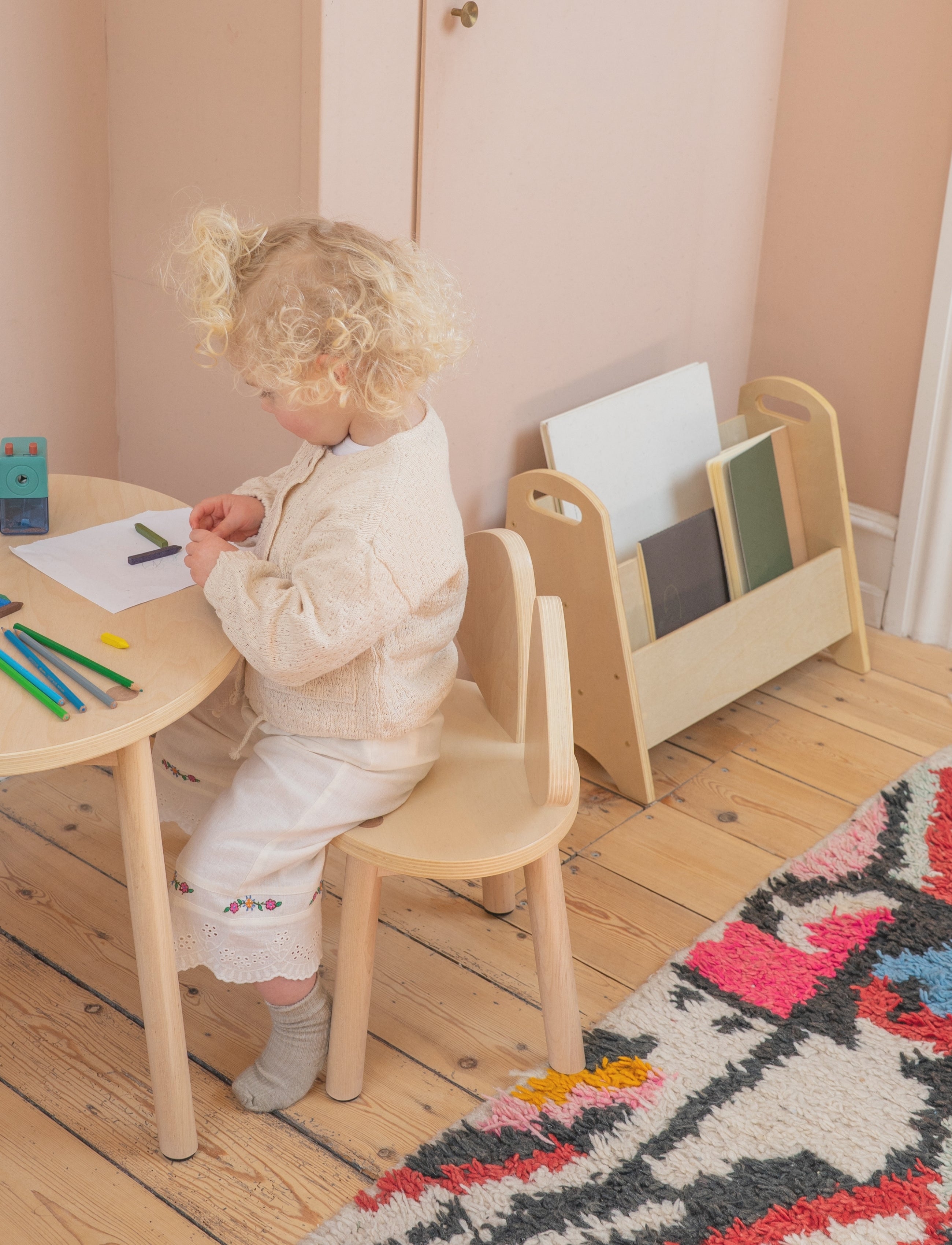 A toddler with curly blonde hair sits at a small round wooden table, drawing on paper. The child wears a cream knit cardigan and cream trousers, sitting on a matching light wood chair.
