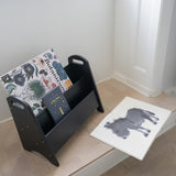 Black magazine rack with books on a wooden floor next to a white wall.