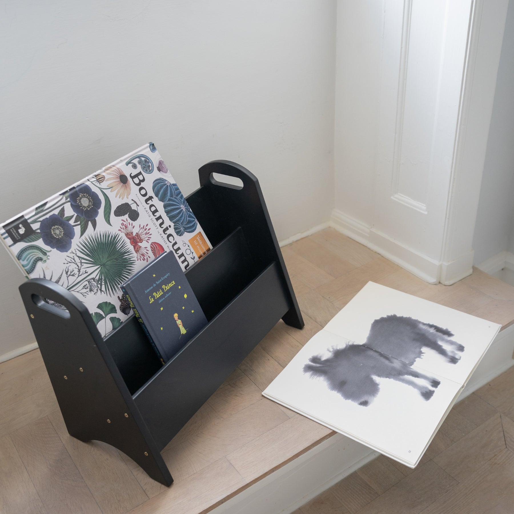 Black magazine rack with books on a wooden floor next to a white wall.