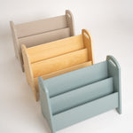 Three wooden magazine racks in different colors: beige, natural wood, and light blue, arranged diagonally on a white background.