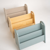 Three wooden magazine racks in different colors: beige, natural wood, and light blue, arranged diagonally on a white background.